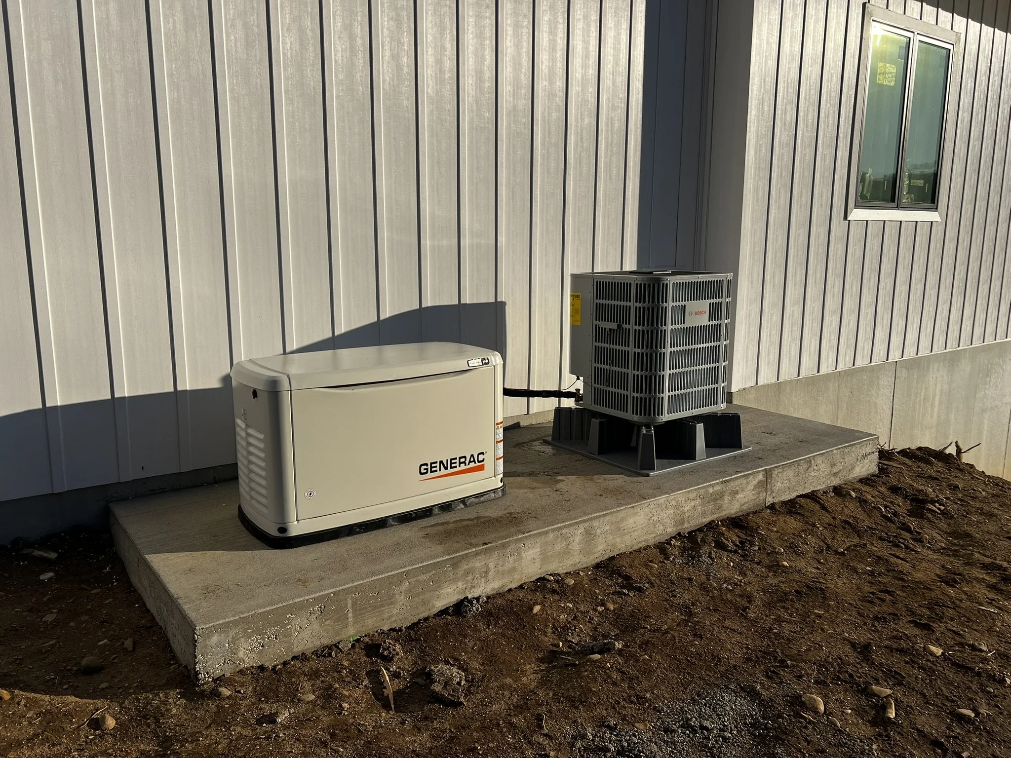 An outdoor scene showing a beige Generac generator and an air conditioning unit installed on a concrete pad against the exterior wall of a building with beige vertical siding and a small window.