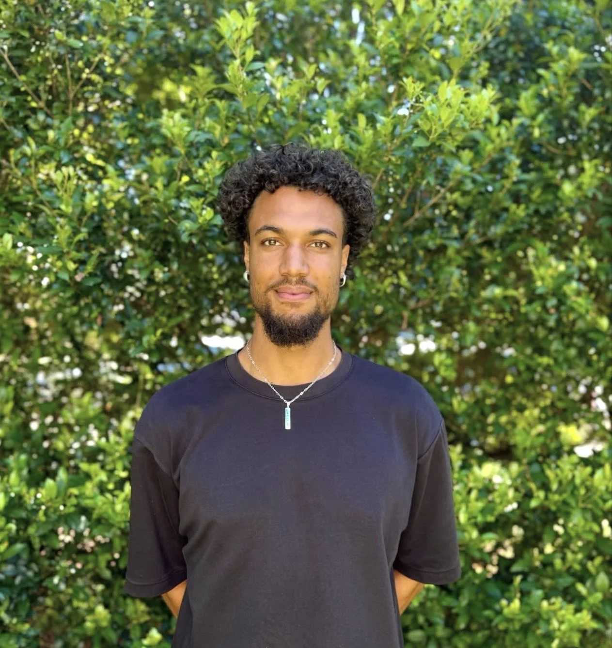 Portrait of a young man with dark, curly hair and a beard, wearing a black t-shirt and a silver necklace, standing outdoors in front of lush green foliage.