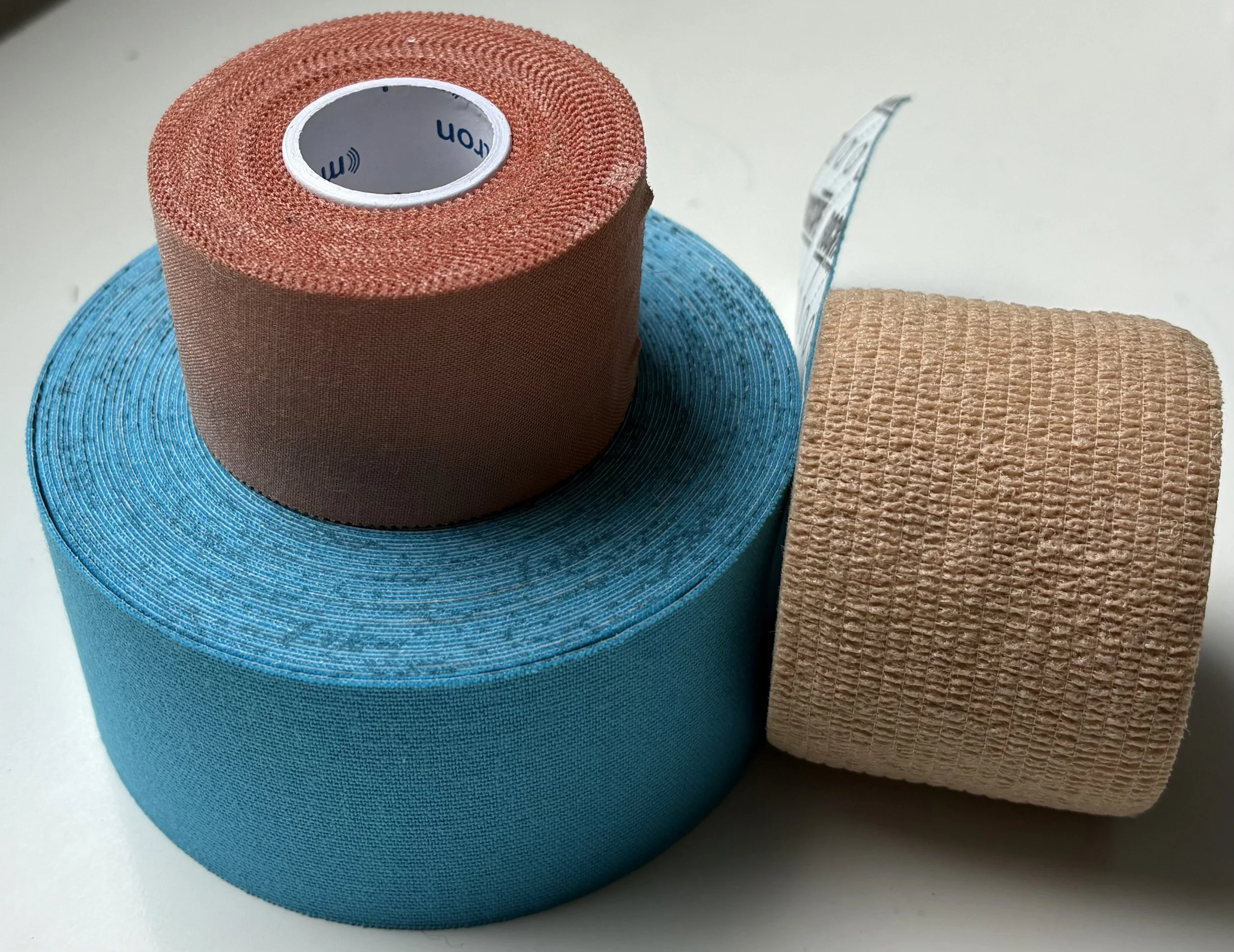 Three rolls of kinesiology or athletic tape in brown, blue, and beige, stacked and arranged on a white surface.
