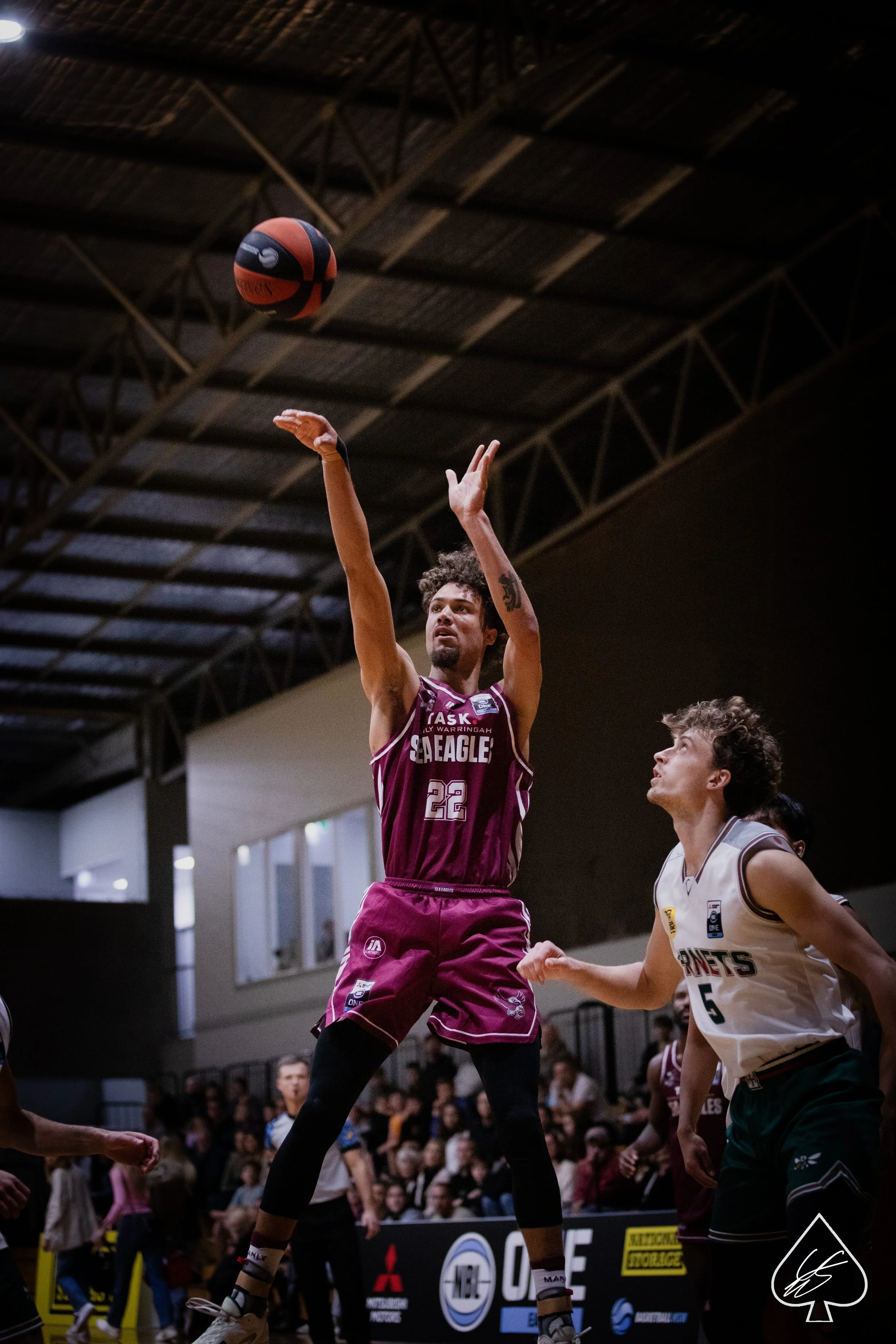 A basketball player wearing a maroon uniform with the number 22 jumps to shoot the ball during a game. Another player in a white uniform with the number 5 is nearby, watching the play.