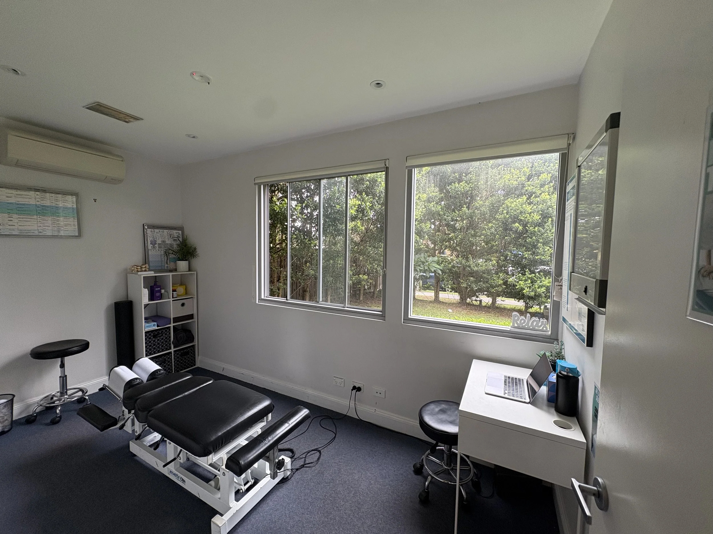 Medical examination room with an adjustable bed, a white desk with a laptop, a stool, a small shelving unit, a whiteboard, and large windows showing greenery outside.