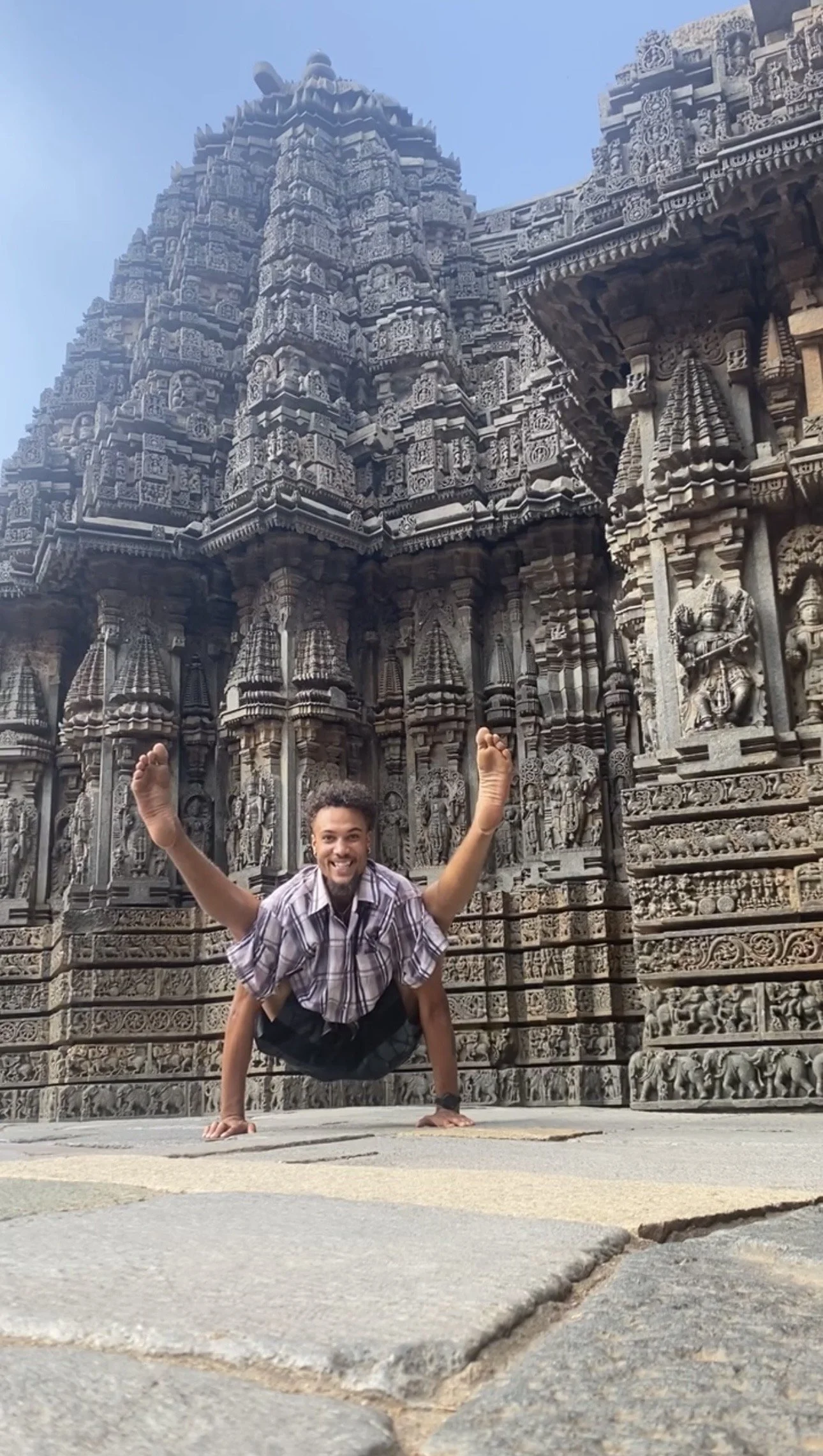 Man performing a yoga pose on the ground in front of an intricately carved stone temple with detailed sculptures.