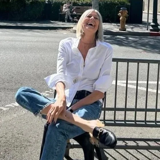 A woman styled in designer clothing wearing a white blouse and jeans. Wearing Gucci slides.