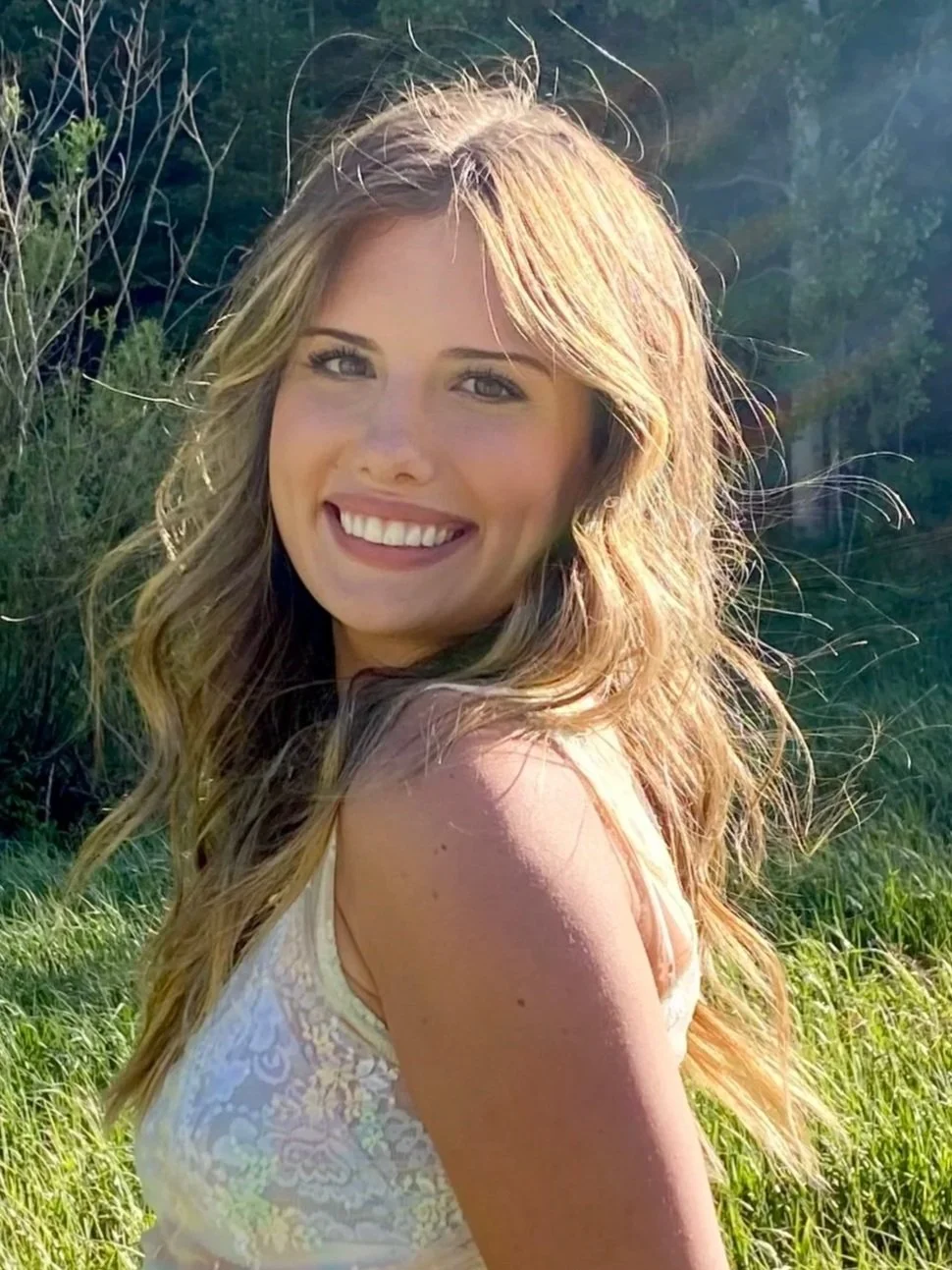 A woman with long blonde hair smiling outdoors in a grassy area with trees in the background.