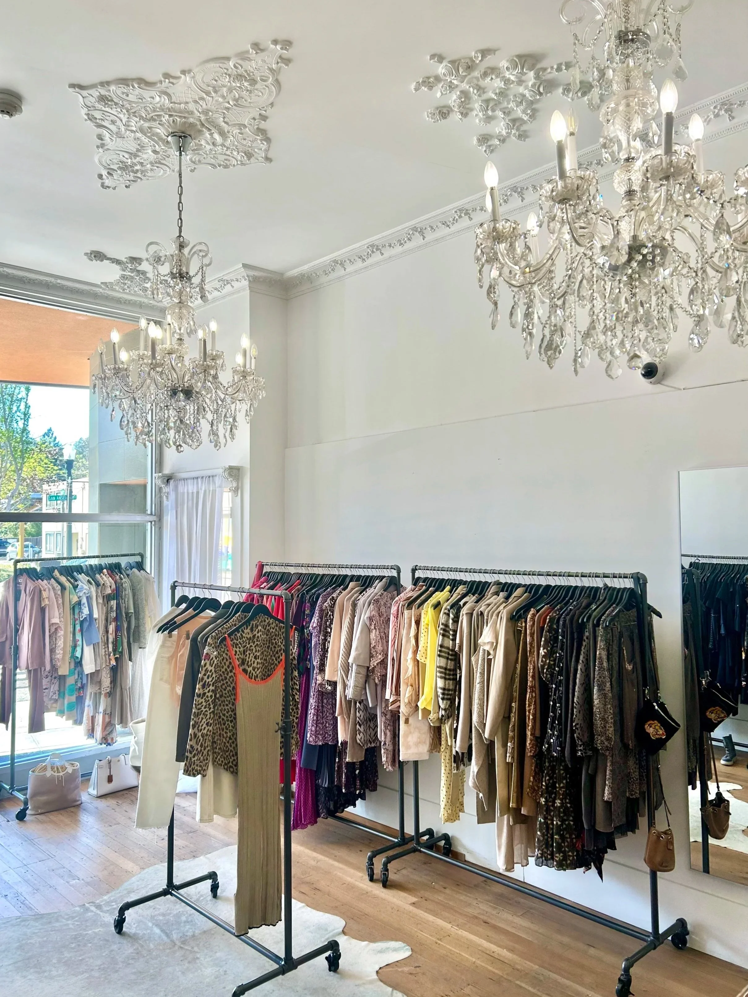 Clothing store interior with racks of color blocked clothes, chandeliers on white ceiling, and a large window with natural light.
