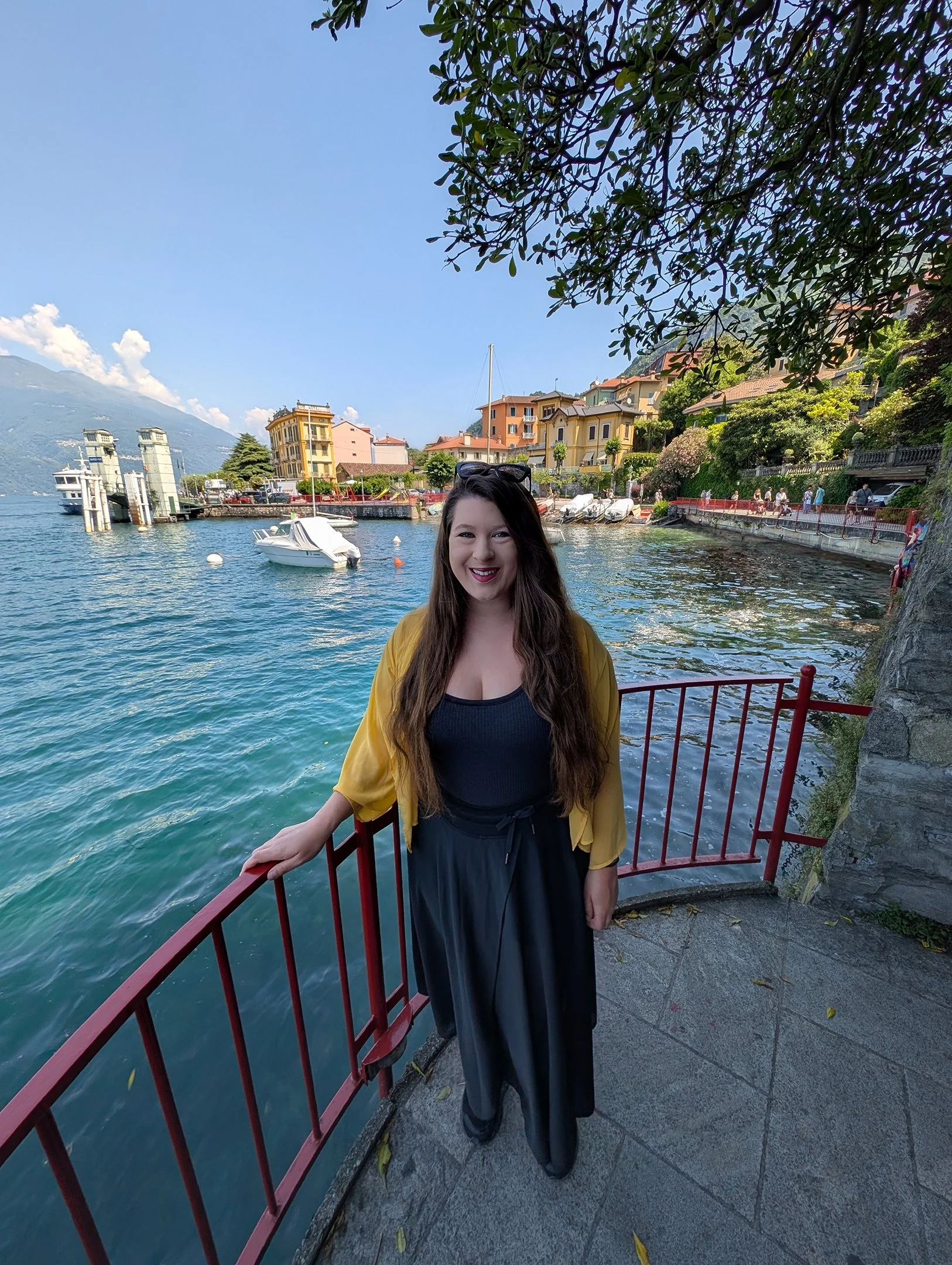 A woman in a black dress and yellow jacket smiling while standing near a red railing by a lake or canal, with boats and colorful buildings in the background, under a clear blue sky.