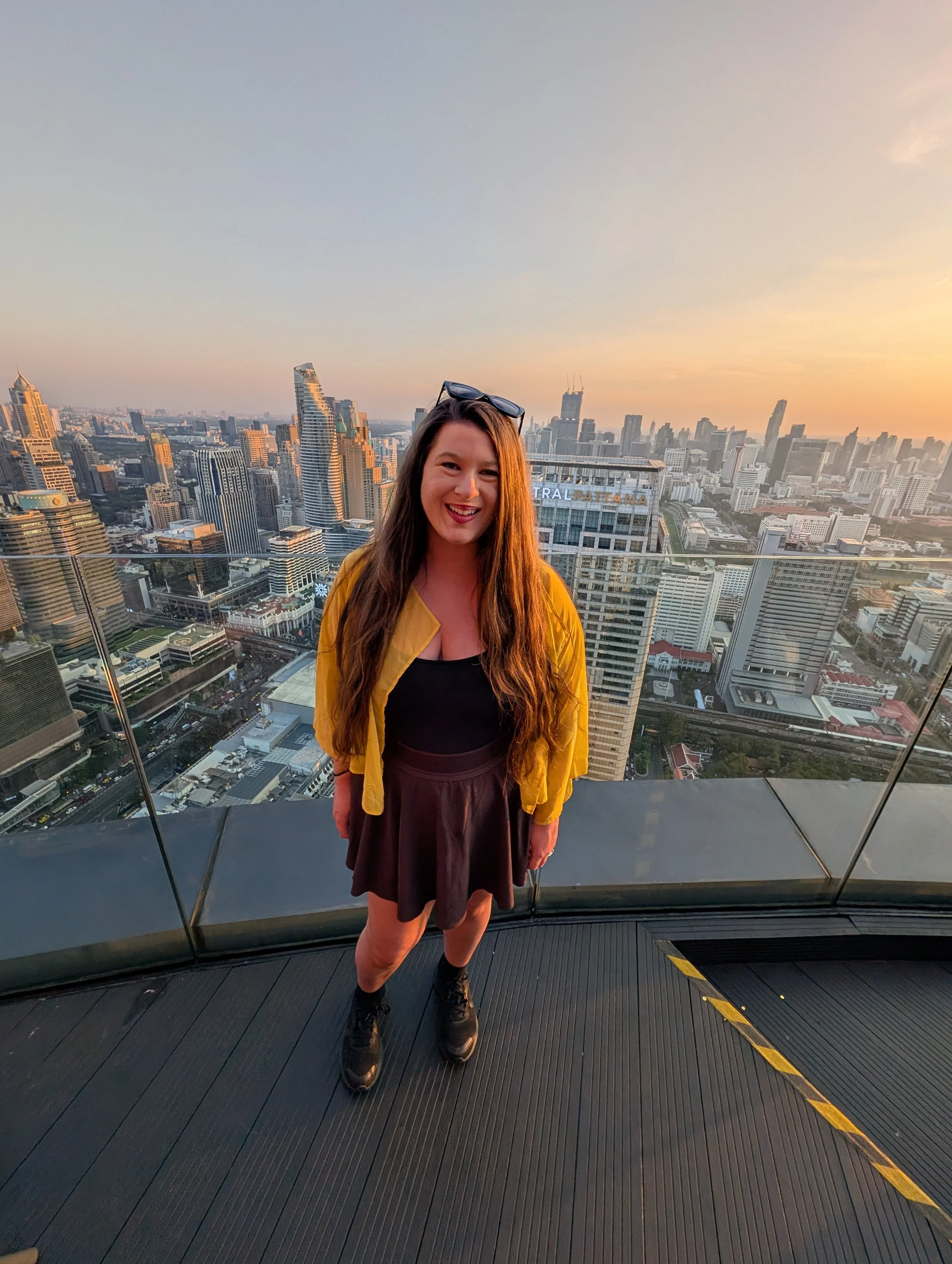 Woman with long brown hair wearing a yellow jacket, black top, and black skirt, standing on a rooftop with a city skyline in the background during sunset.