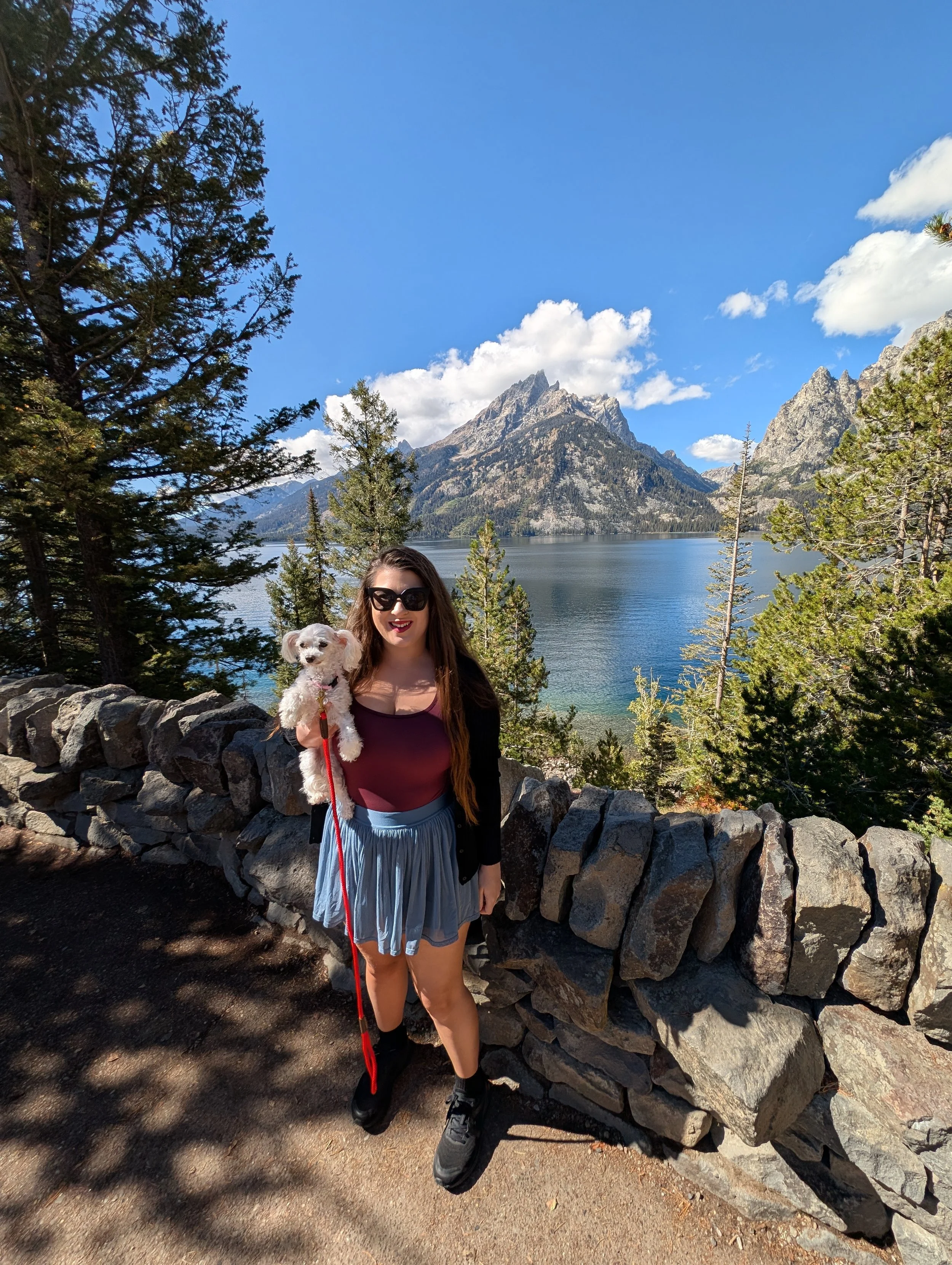 A woman holding a small white dog stands in front of a stone fence with a lake, trees, and mountains in the background on a sunny day.