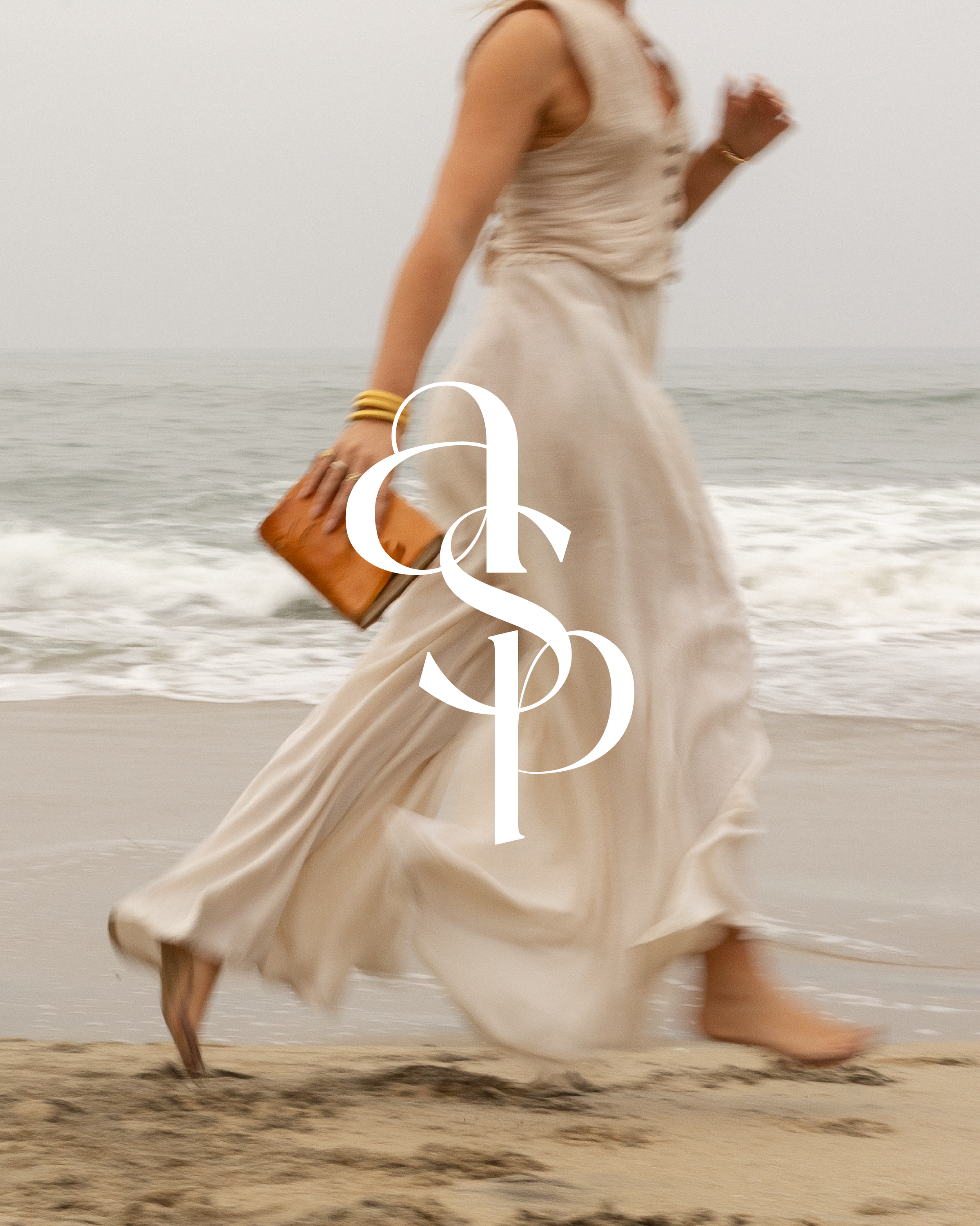 A woman walking barefoot on the beach wearing a flowing cream-colored dress, carrying a brown clutch, with the ocean in the background and a large white logo overlay reading 'asp'.}