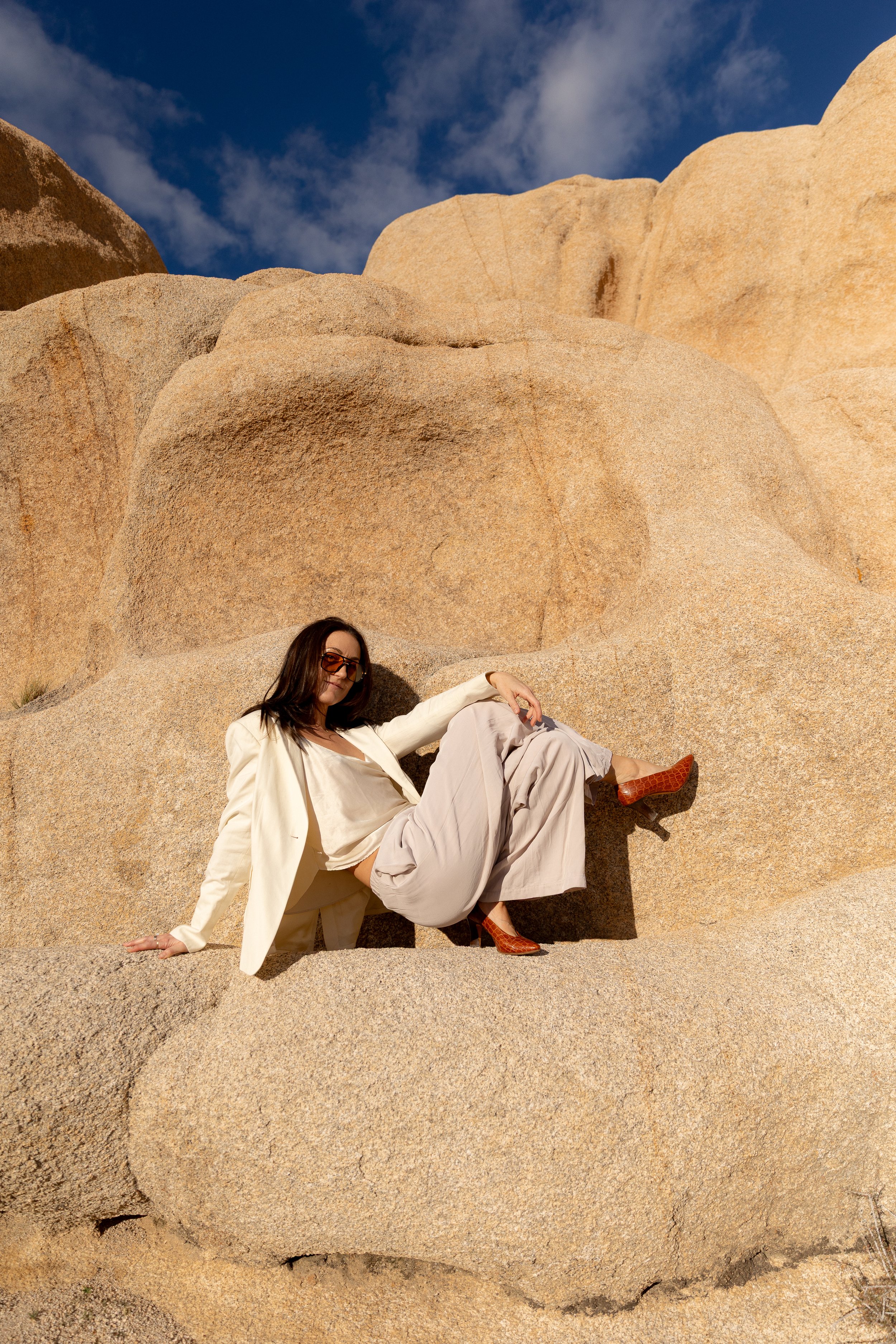 A woman in a cream suit and beige trousers, wearing sunglasses and brown heels, sitting on large rocks in a desert environment with a blue sky and clouds in the background.