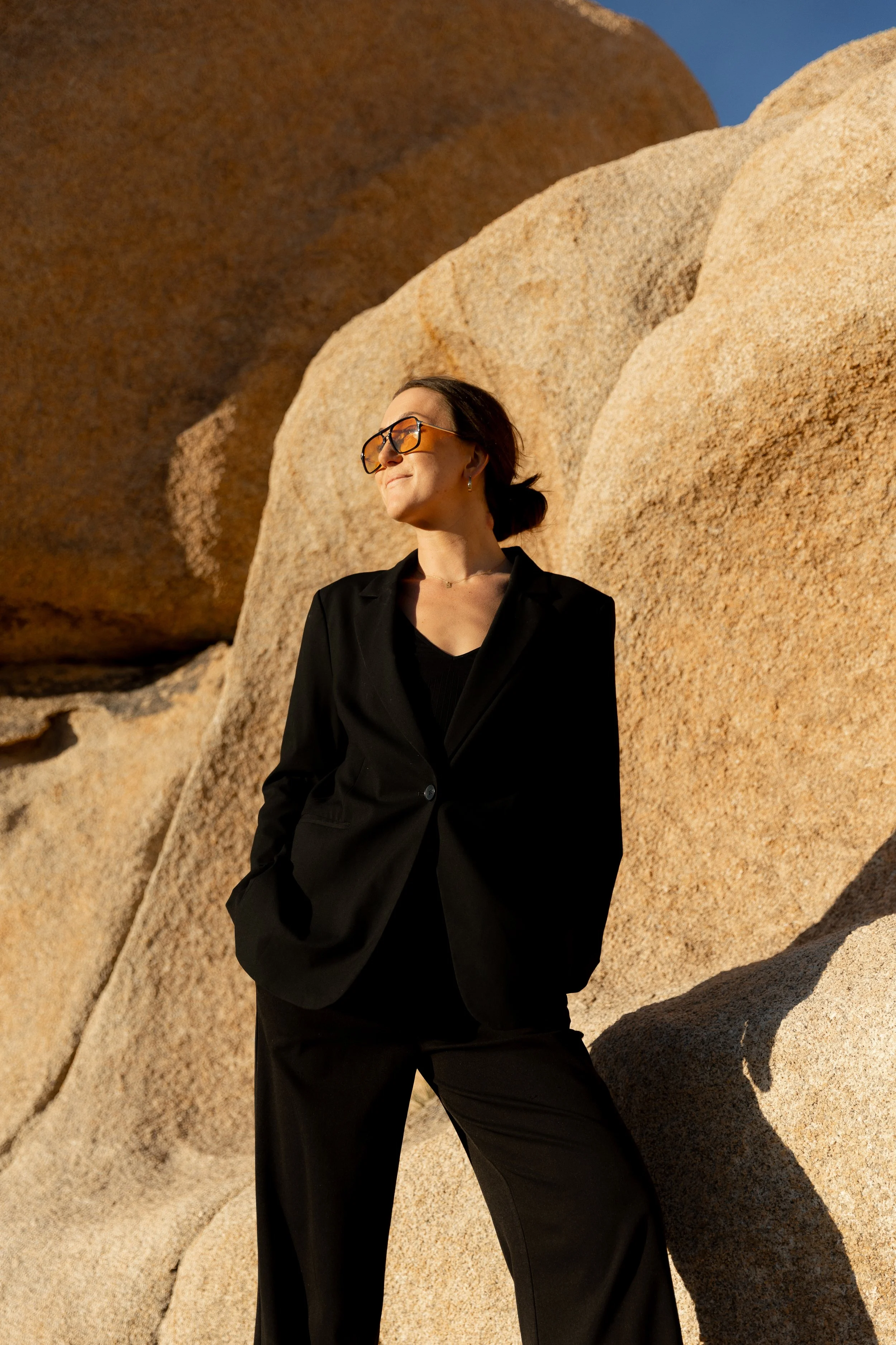 A woman in a black suit with sunglasses standing outdoors against large rocks.