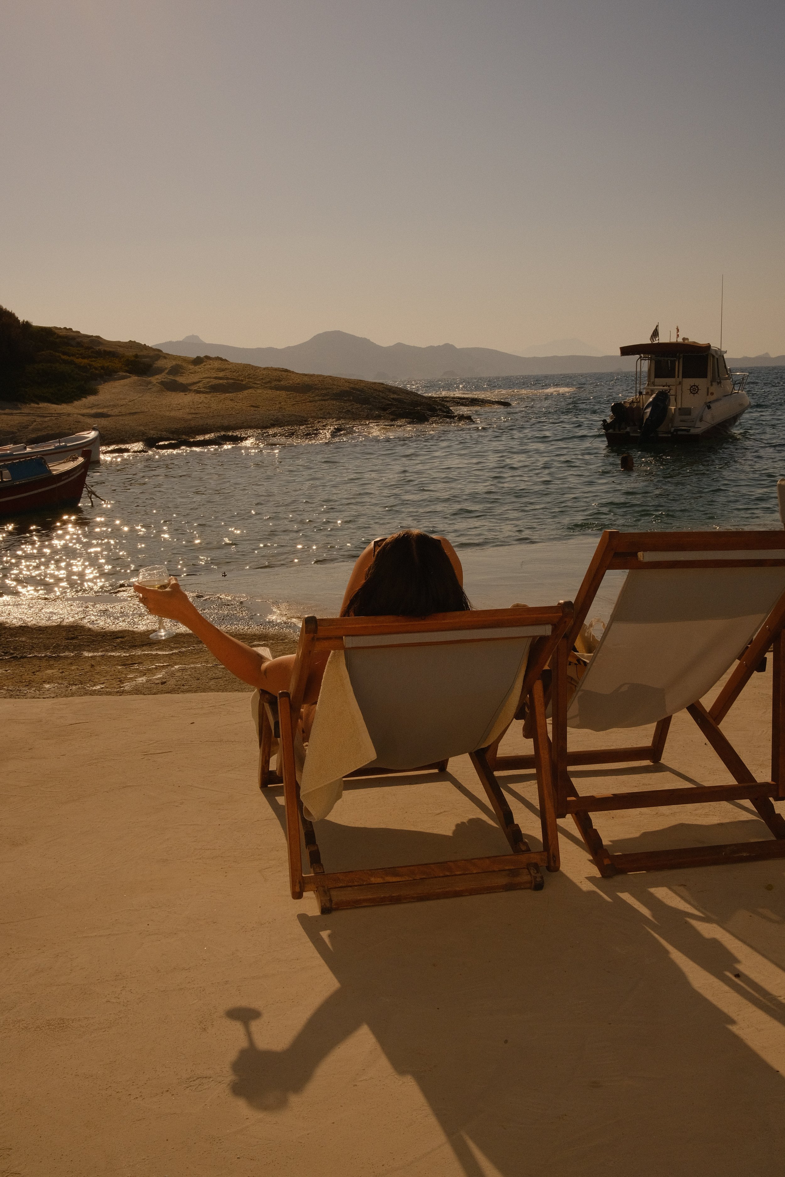 A woman relaxing on a lounge chair by the water, holding a drink, with boats and a rocky coastline in the background.