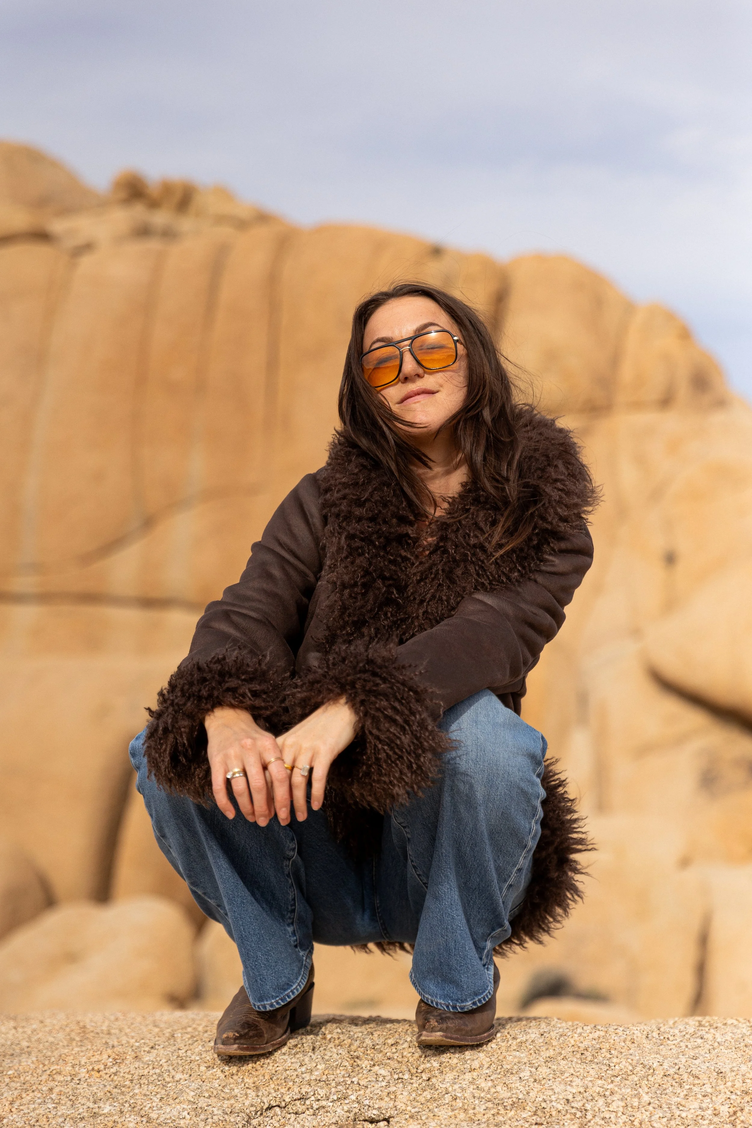 A woman crouching on a sandy surface with large beige rock formations behind her, wearing sunglasses, a brown fur-lined coat, blue jeans, and brown boots.