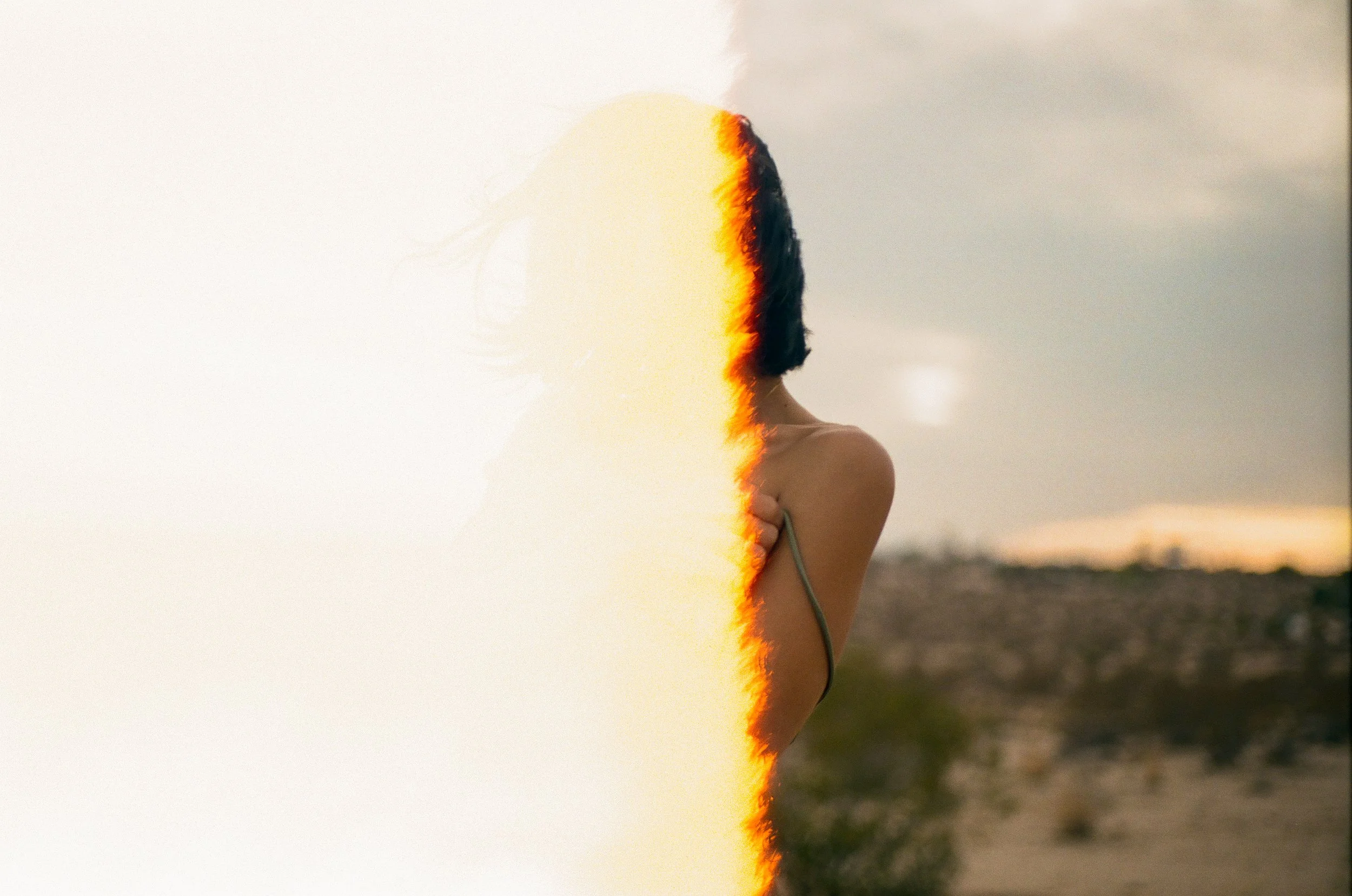 Half of a woman with dark hair and a bare shoulder, standing outdoors during sunset, with the other half showing digital distortion and a burnt edge effect.