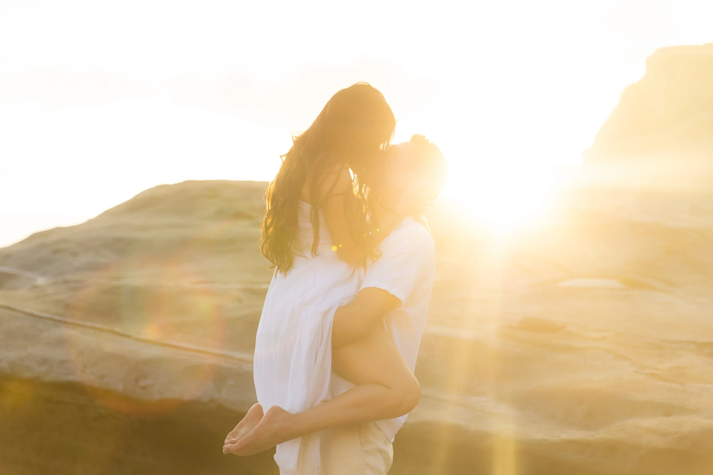A person lifting a woman in front of a sunset on a desert landscape.