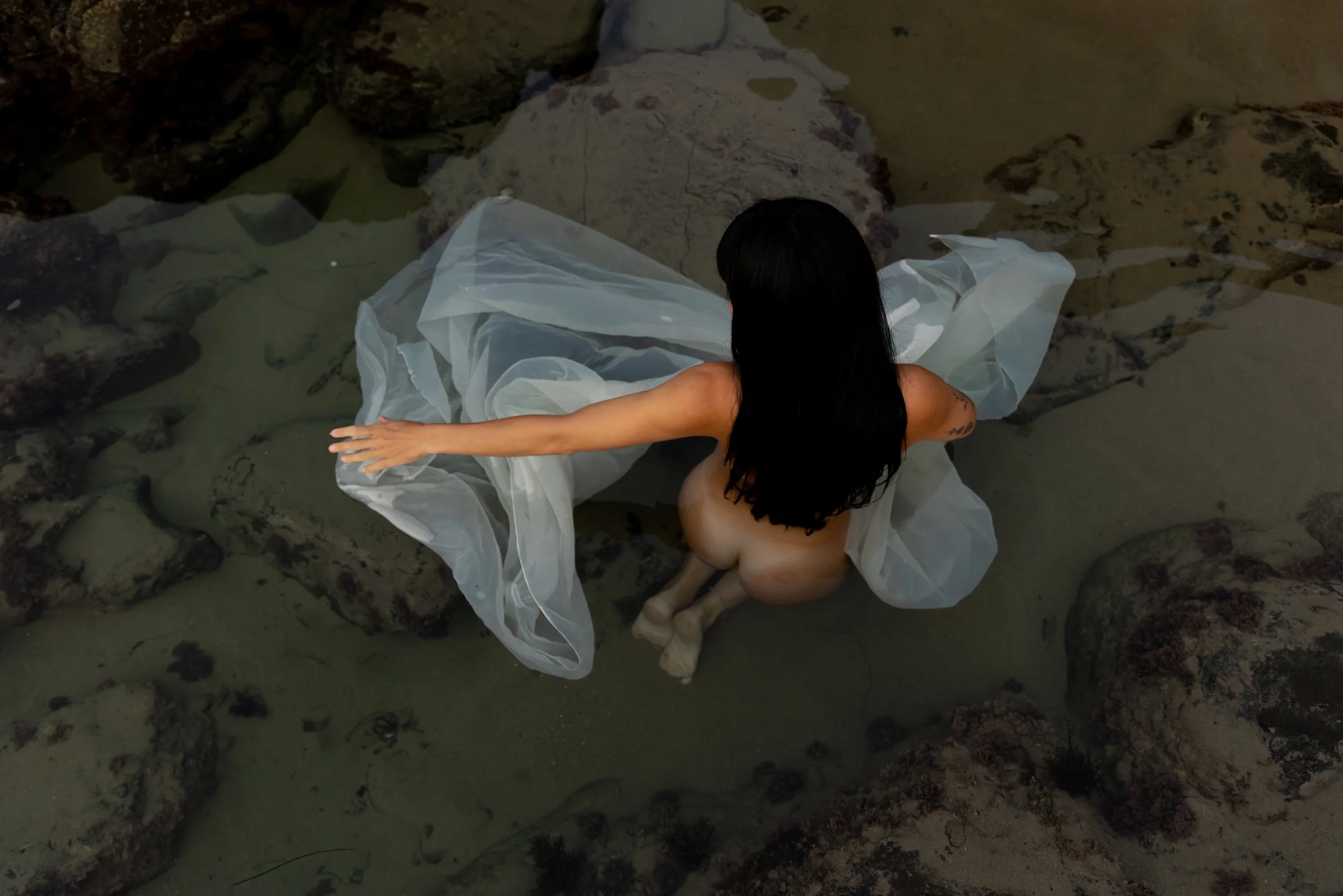 A woman with long dark hair kneeling naked in shallow water, holding a sheer white fabric while surrounded by rocks.