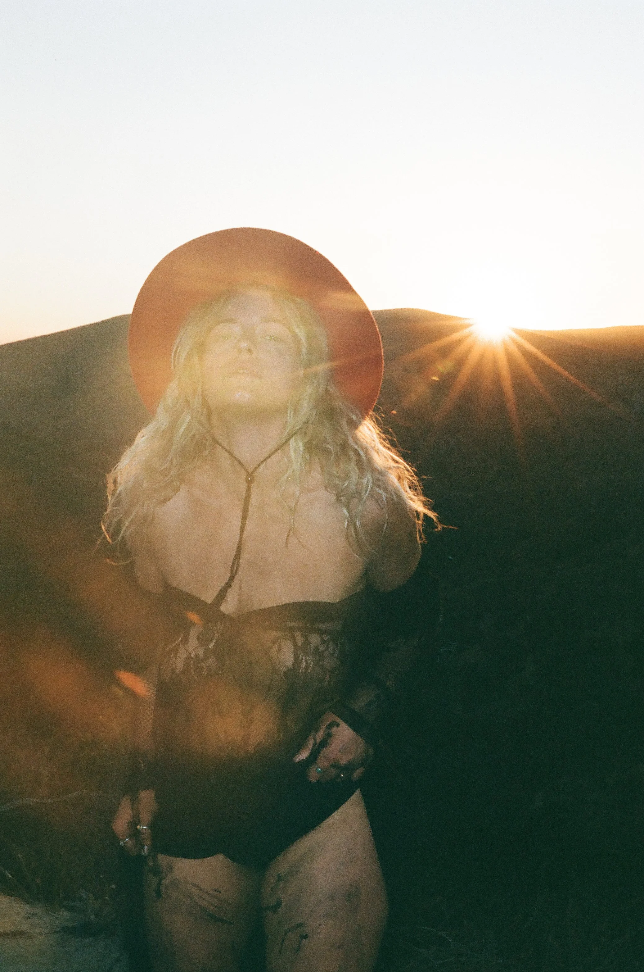 A woman in a black lace top and shorts standing outdoors with the sun setting behind hills, wearing a wide-brimmed hat and looking at the camera.