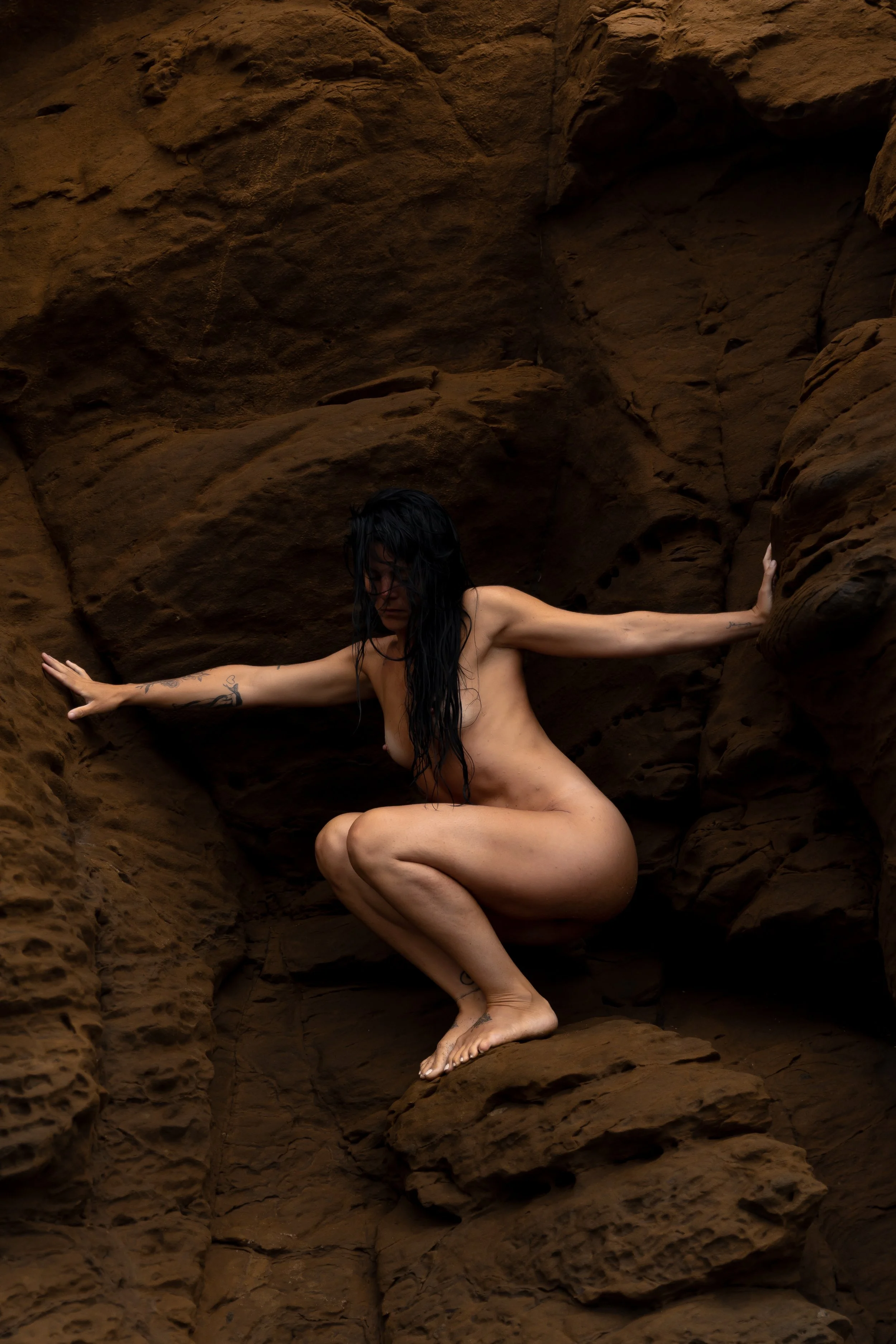A nude woman with wet black hair crouching between reddish-brown rock formations, with her arms extended to the sides touching the rocks.