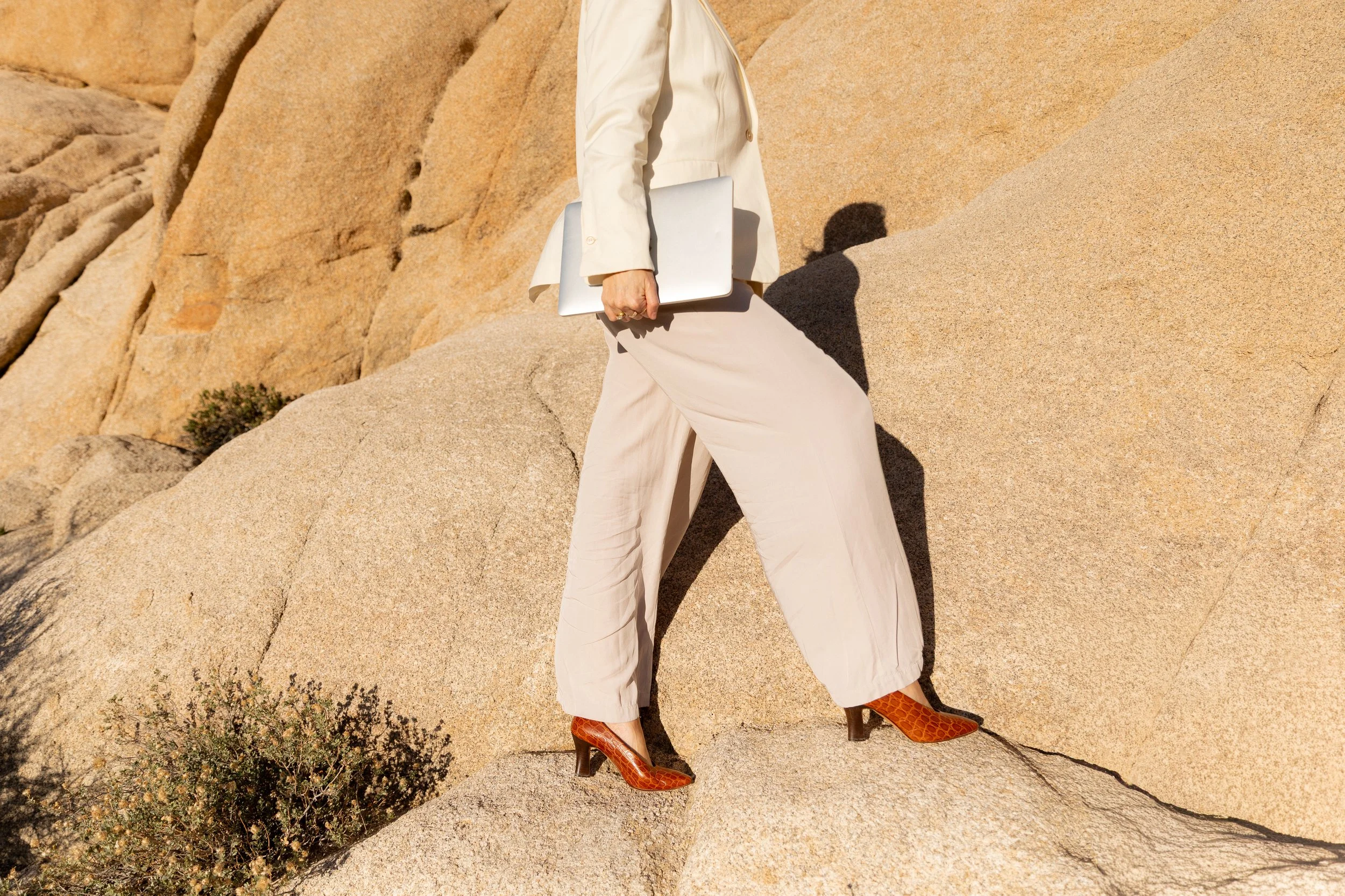 Person wearing cream-colored suit and orange high-heeled boots standing on rocky terrain and holding a silver laptop.