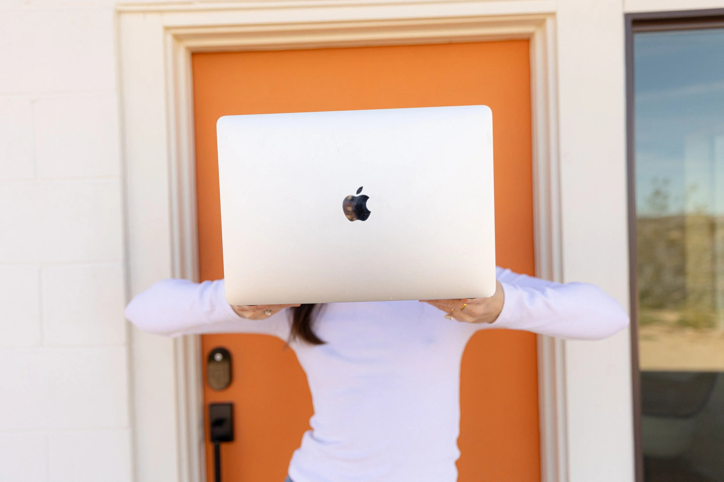 Person holding a silver Apple MacBook laptop in front of an orange door, obscuring their face, wearing a white long-sleeve shirt.