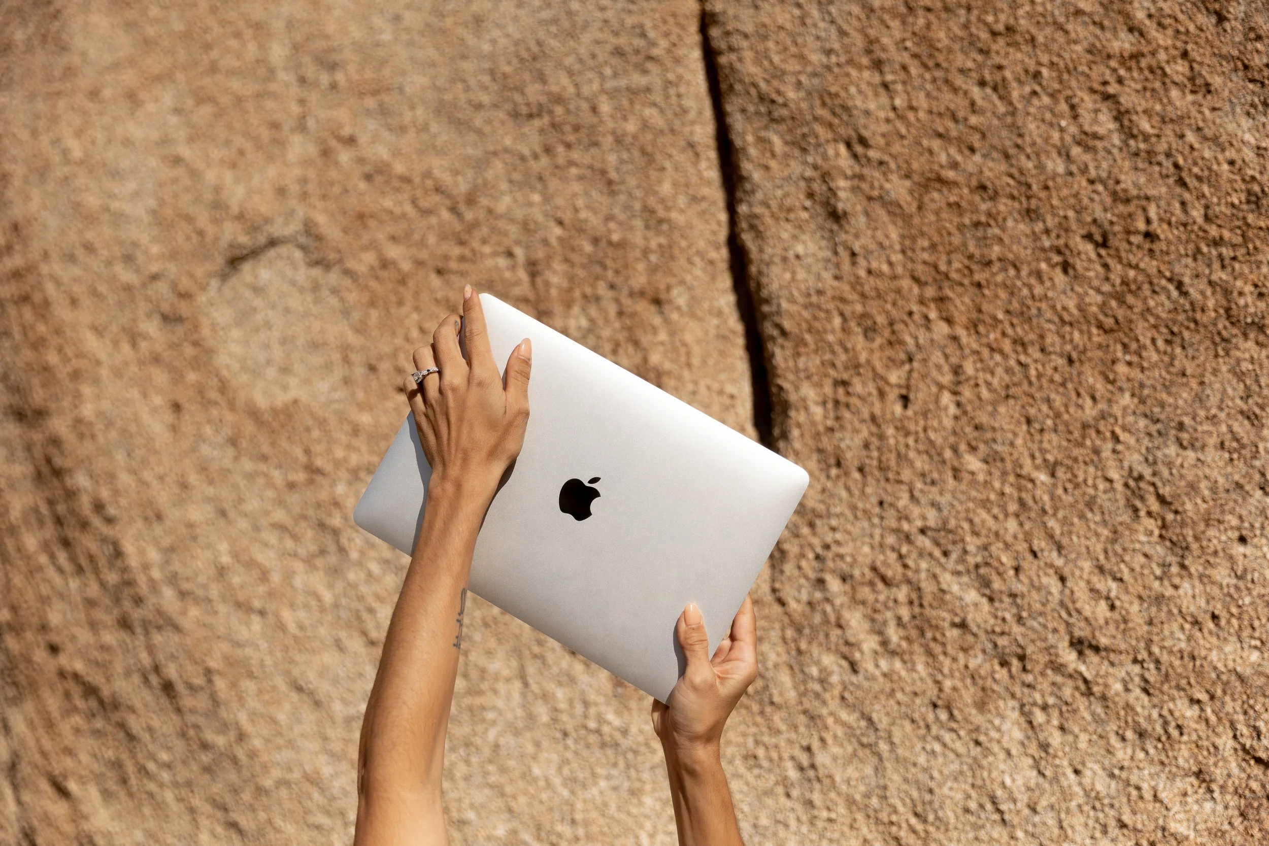 A person holding a silver MacBook with an Apple logo, against a textured rock wall background.
