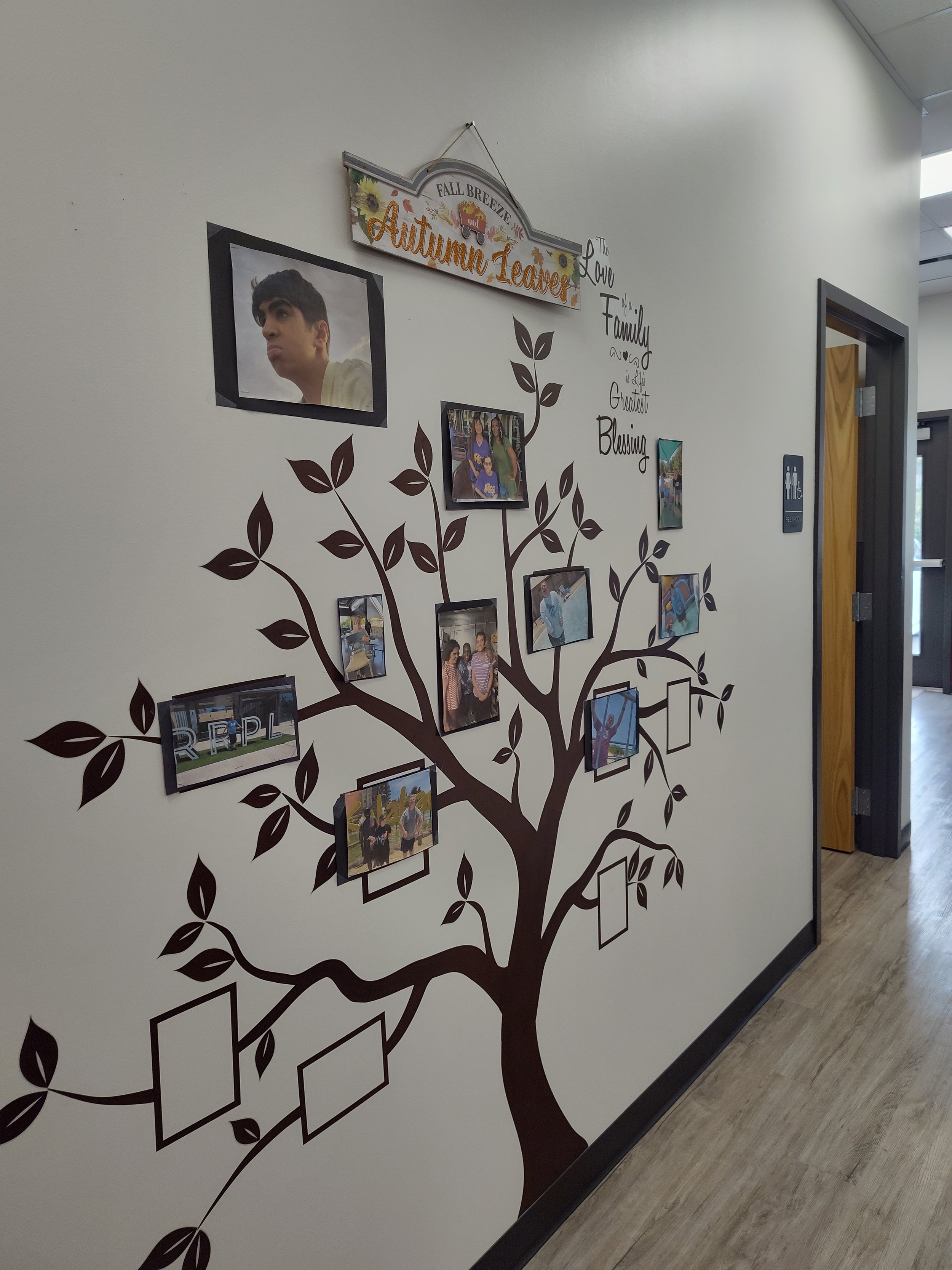 Decorative wall with a tree design, photos, and frames, featuring a sign that reads 'Fall Breeze Autumn Leaves.' Text on the wall says 'The Love & Family... and the Greatest Blessing.'