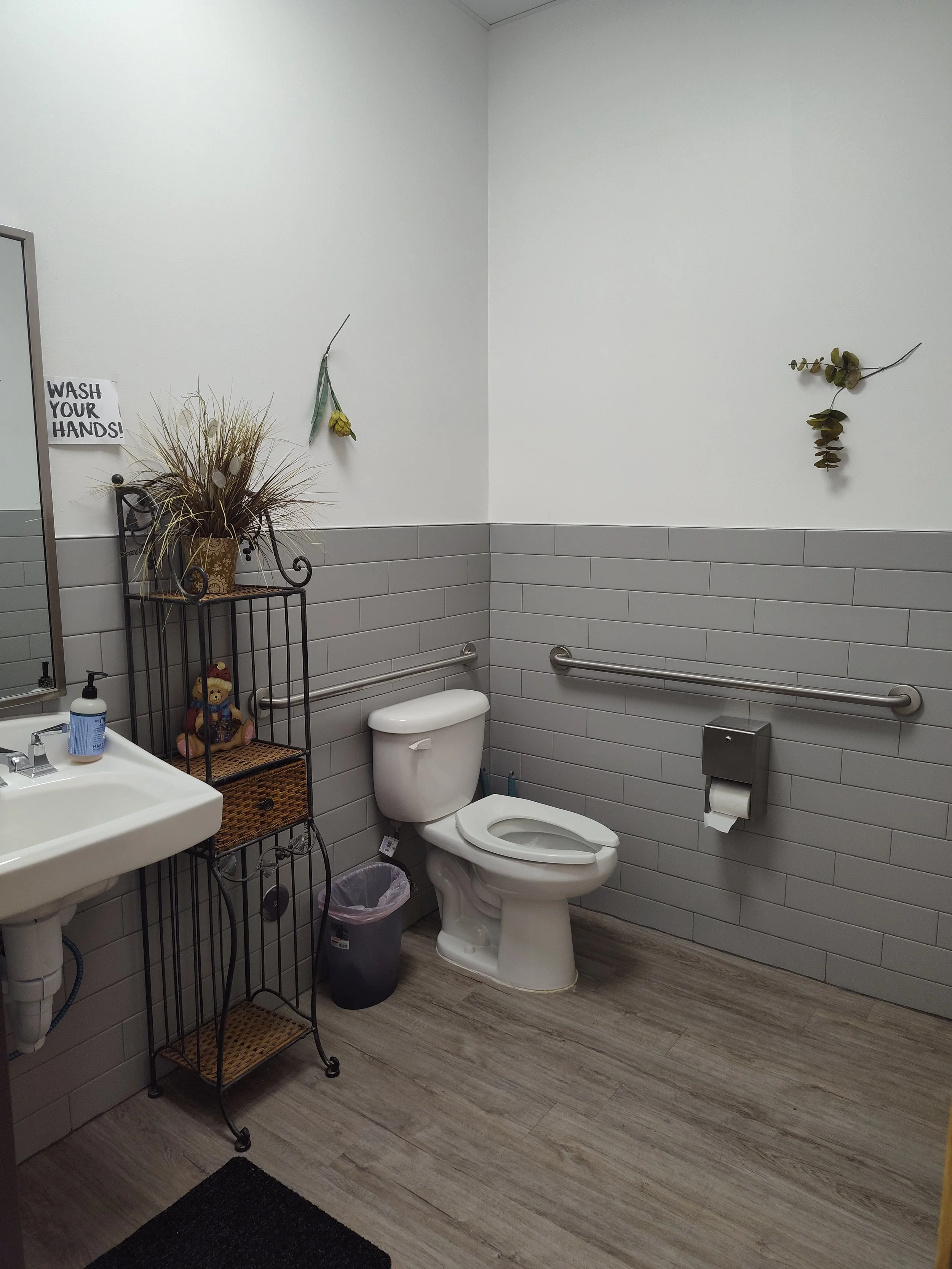Accessible restroom with gray tiled walls, a white toilet, a small sink with soap dispenser, a black metal shelf with a teddy bear, decorative dried plants, and a sign that says 'Wash Your Hands!'