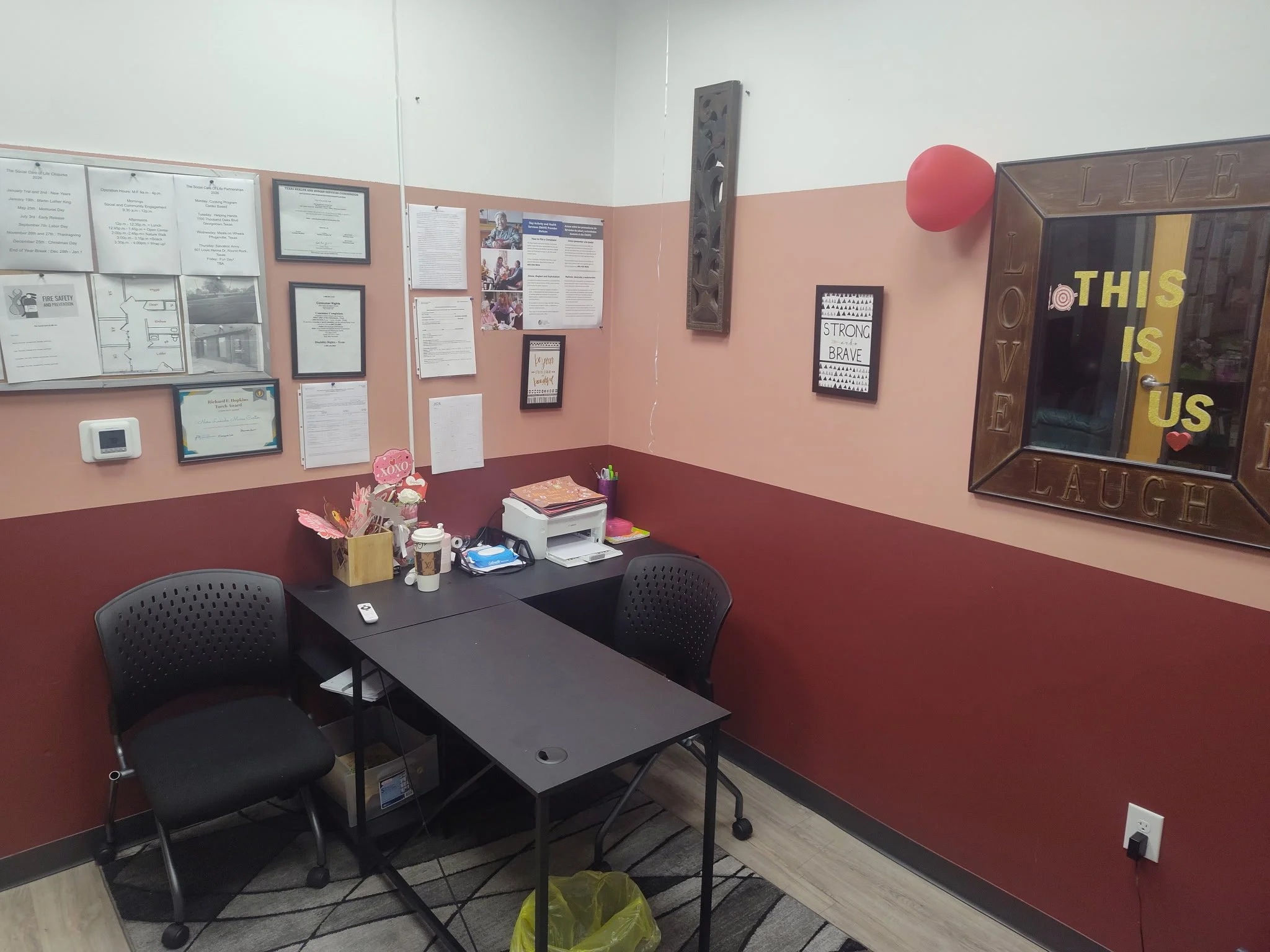 A foyer space with two black chairs, a black table, a wall decorated with various framed documents and certificates, a bulletin board, and motivational signs. There is a small mirror and the words 'This is us' in yellow on the mirror. 