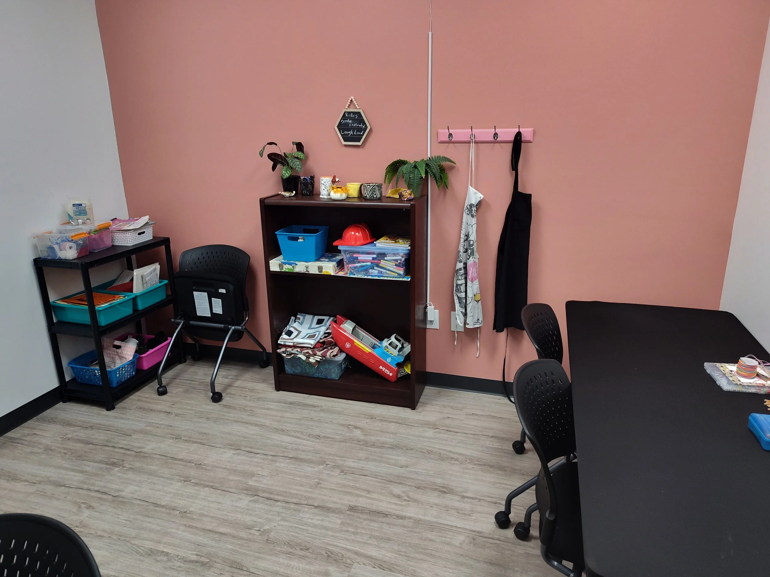 A small classroom or craft room with black tables, black chairs, and a pink accent wall. There are shelves with art supplies, a small black cart with storage bins, and a pink coat rack with hanging aprons or garments.