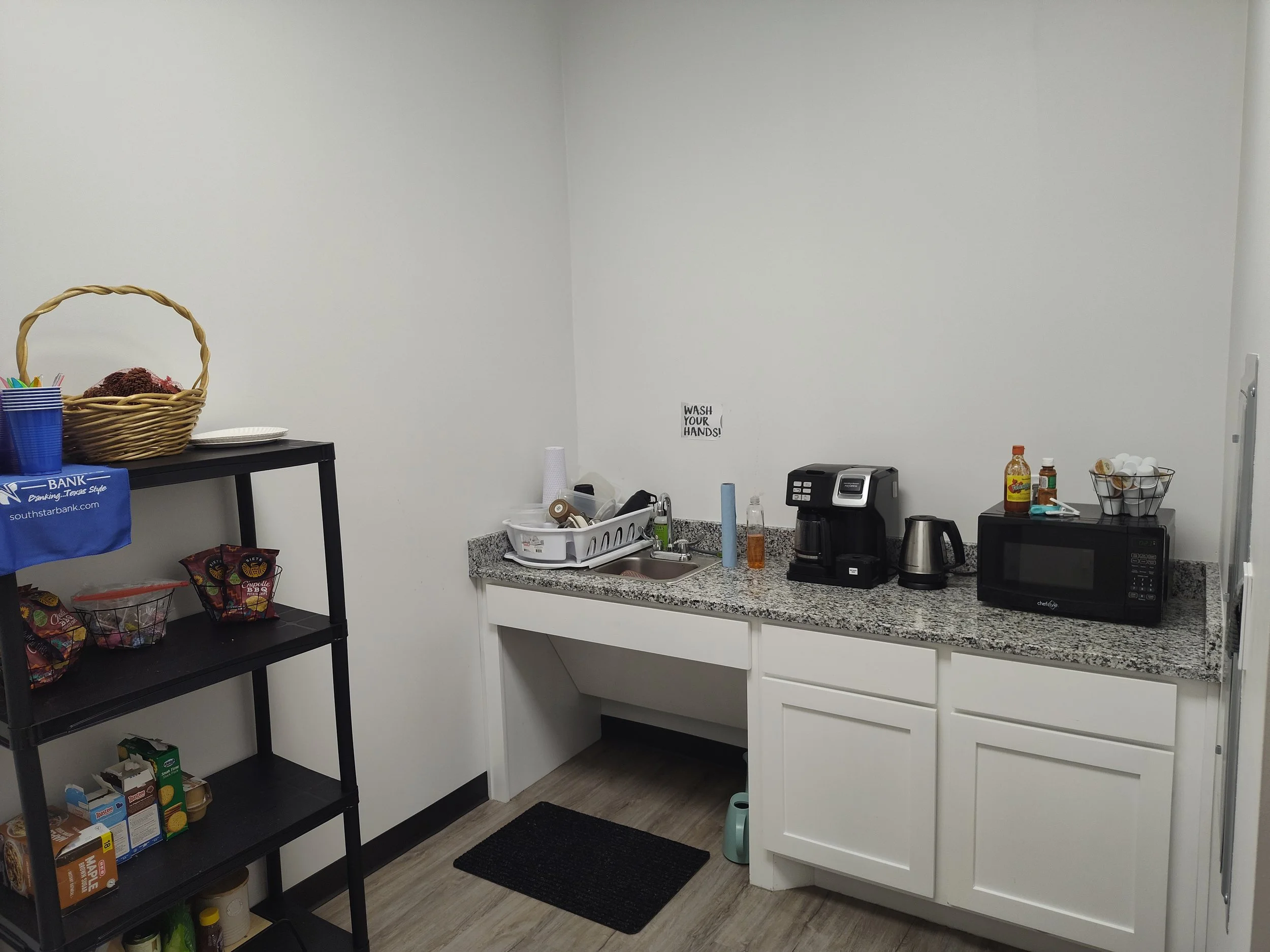 A small kitchenette with white cabinets, gray granite countertop, black coffee maker, electric kettle, microwave, and a sign that says 'Wash Your Hands!'. There is a black shelf with snacks, cups, and paper towels on the left side of the image, and a