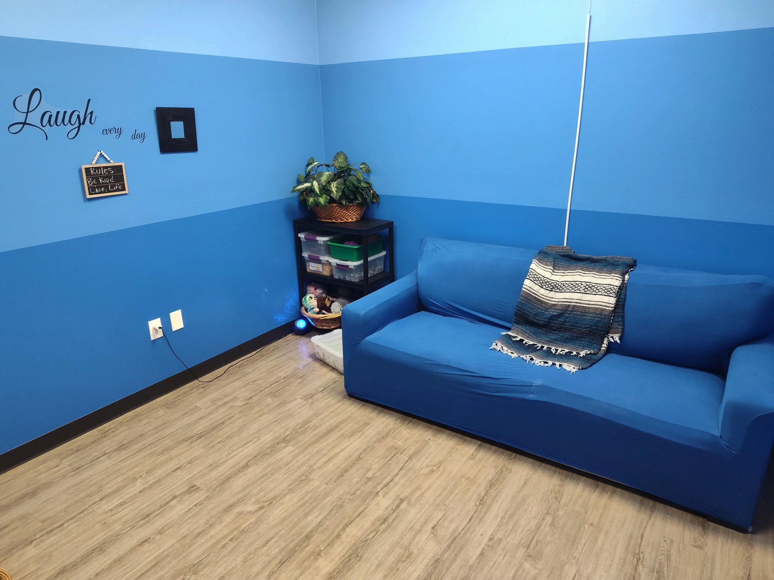 A blue sofa with a multicolored striped throw blanket in a cozy room with blue walls and wooden floor. A small black shelf holding a basket of greenery and storage containers is next to the sofa. Decorative wall signs read "Laugh every day" and a rul