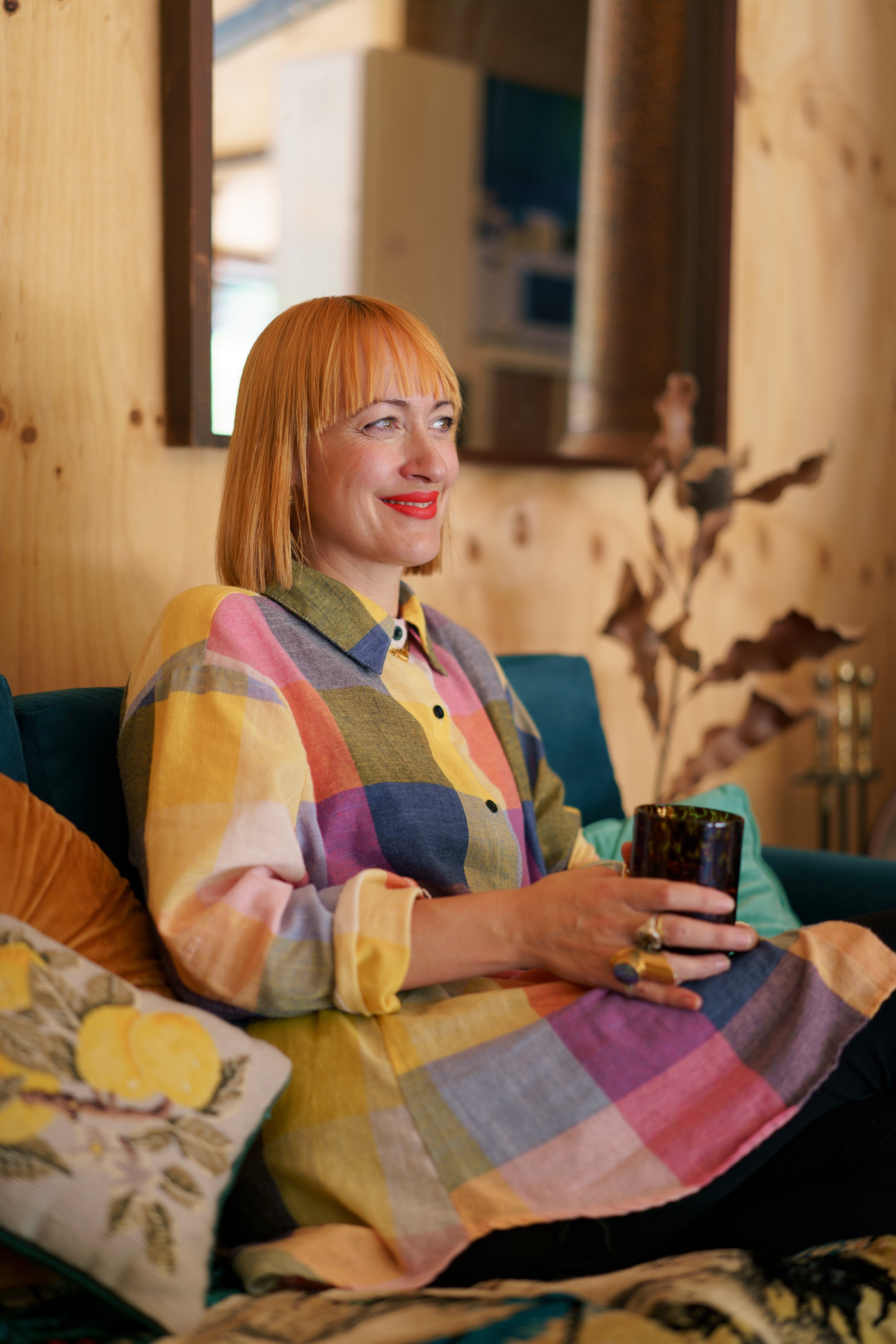 A woman with orange hair, wearing a colorful checkered shirt, sitting on a couch, holding a glass, smiling, in a cozy ceramic workshop studio with wooden walls and a mirror behind her.