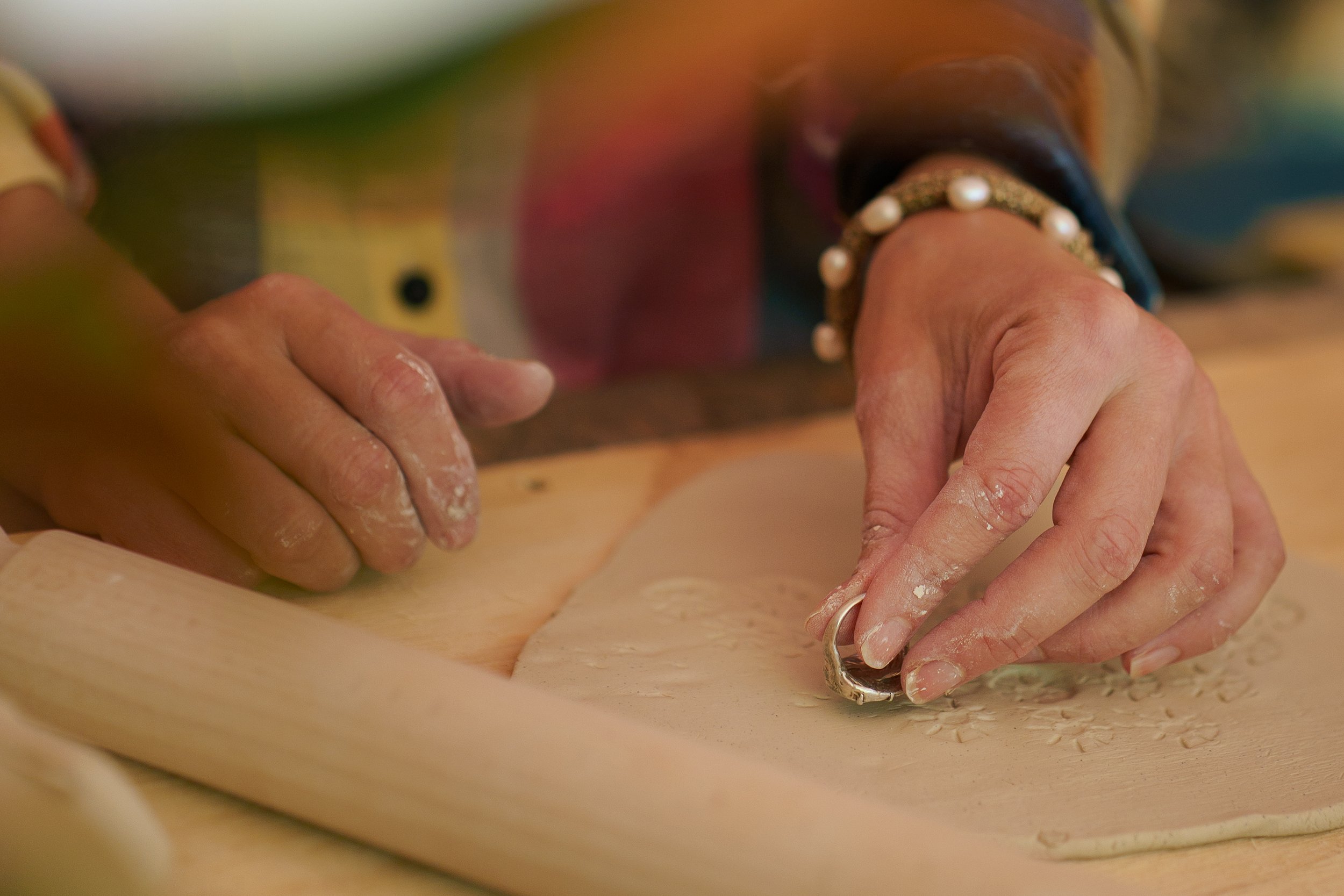 Close-up of hands kneading clay on a wooden workbench, with some green foliage and a bracelet with pearls visible.