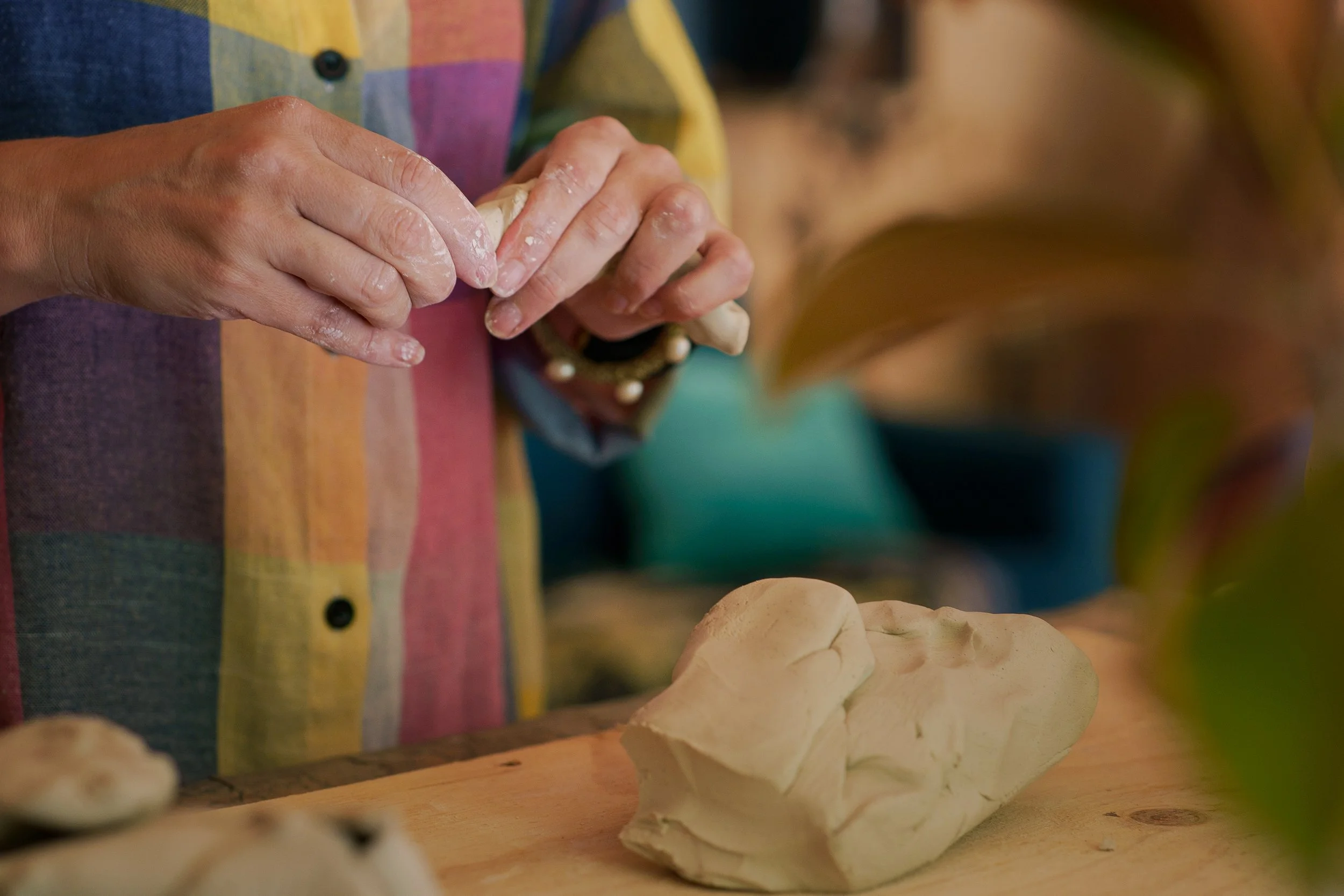 Person mixing clay with hands in a colorful checkered shirt, with rolled clay and rolling pin on a wooden surface.