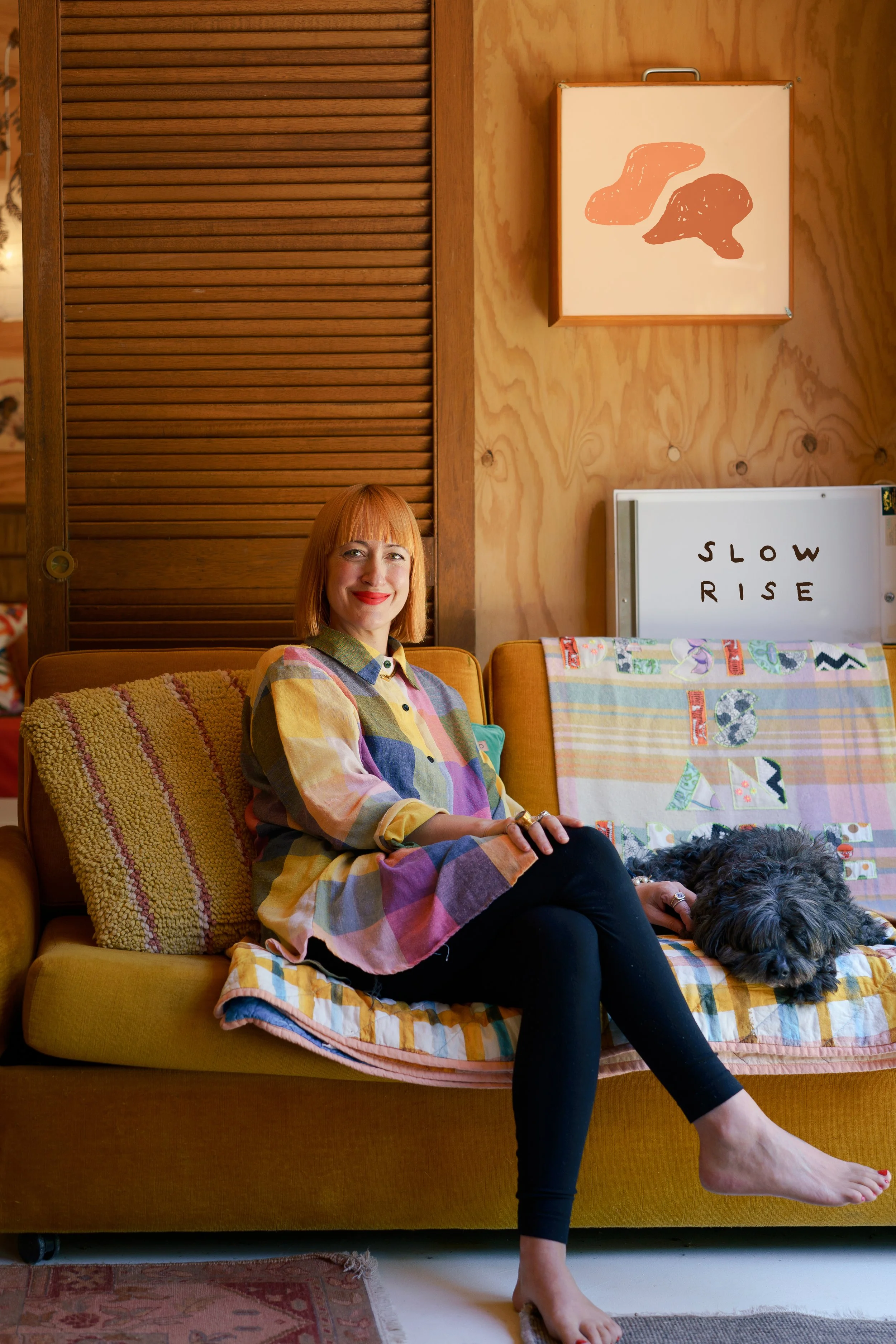 A woman sitting on a yellow sofa, wearing a colorful checkered shirt and black leggings, with a gray fluffy dog lying next to her. The background features wooden wall panels. There is a lightbox with the words 'SLOW RISE' written on it behind her.