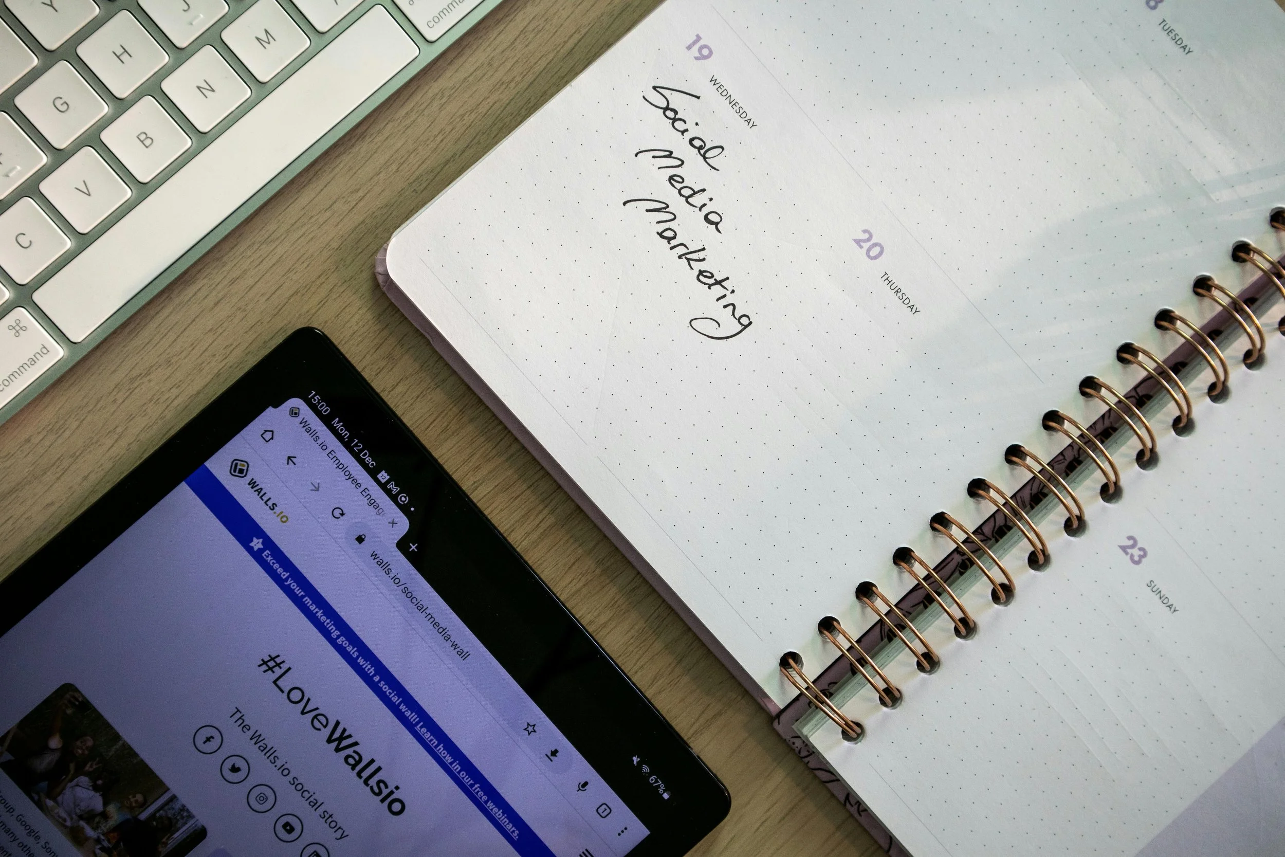 A desk with a white keyboard, a spiral-bound planner open to Wednesday and Thursday pages, a smartphone displaying the Wall's.io website, and a wooden surface.