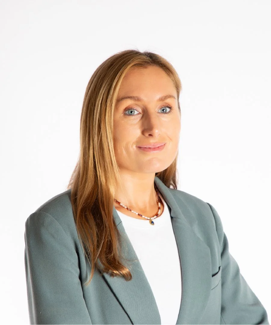 Professional woman with long, light brown hair wearing a gray blazer, white shirt, and a beaded necklace, standing against a plain white background.
