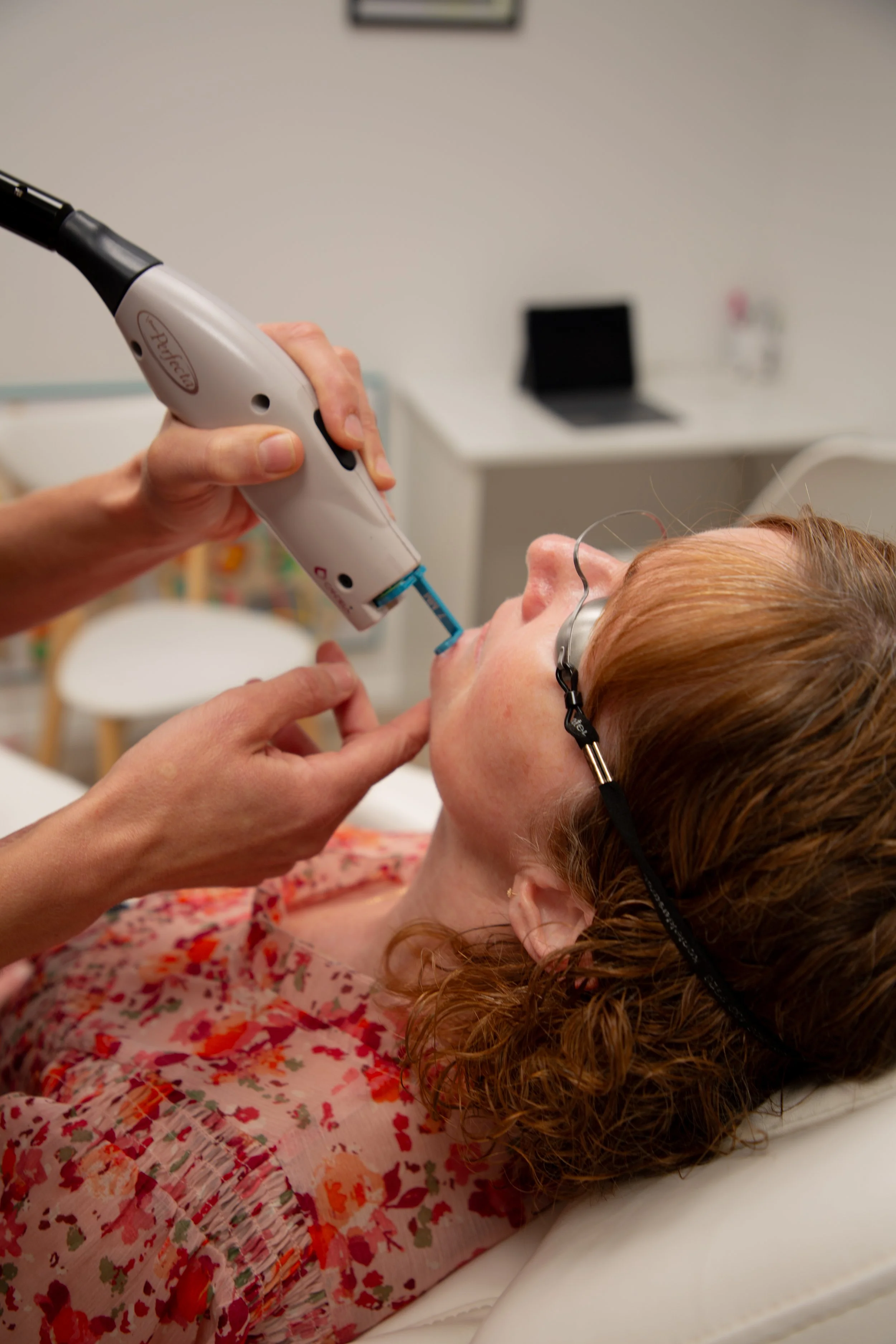 A woman with red hair is lying on a medical bed with eye protection. A healthcare professional is holding a medical device near her mouth, possibly for oral or dental examination.