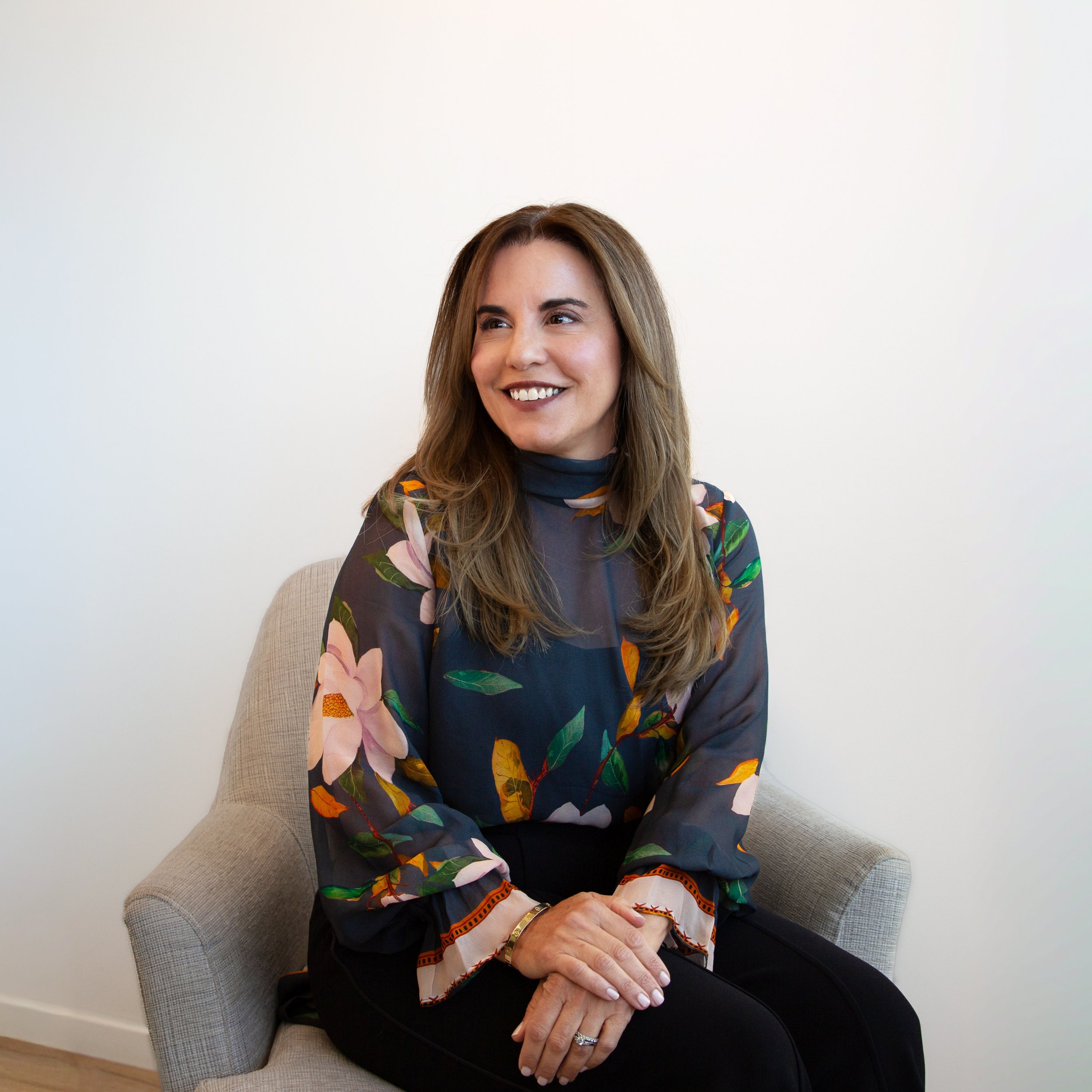 A woman sitting in a beige armchair against a plain white wall, smiling and looking to her right. She has long, wavy brown hair and is wearing a dark shirt with a floral-patterned jacket. Her hands are resting on her lap, with one hand gently grasping the other.