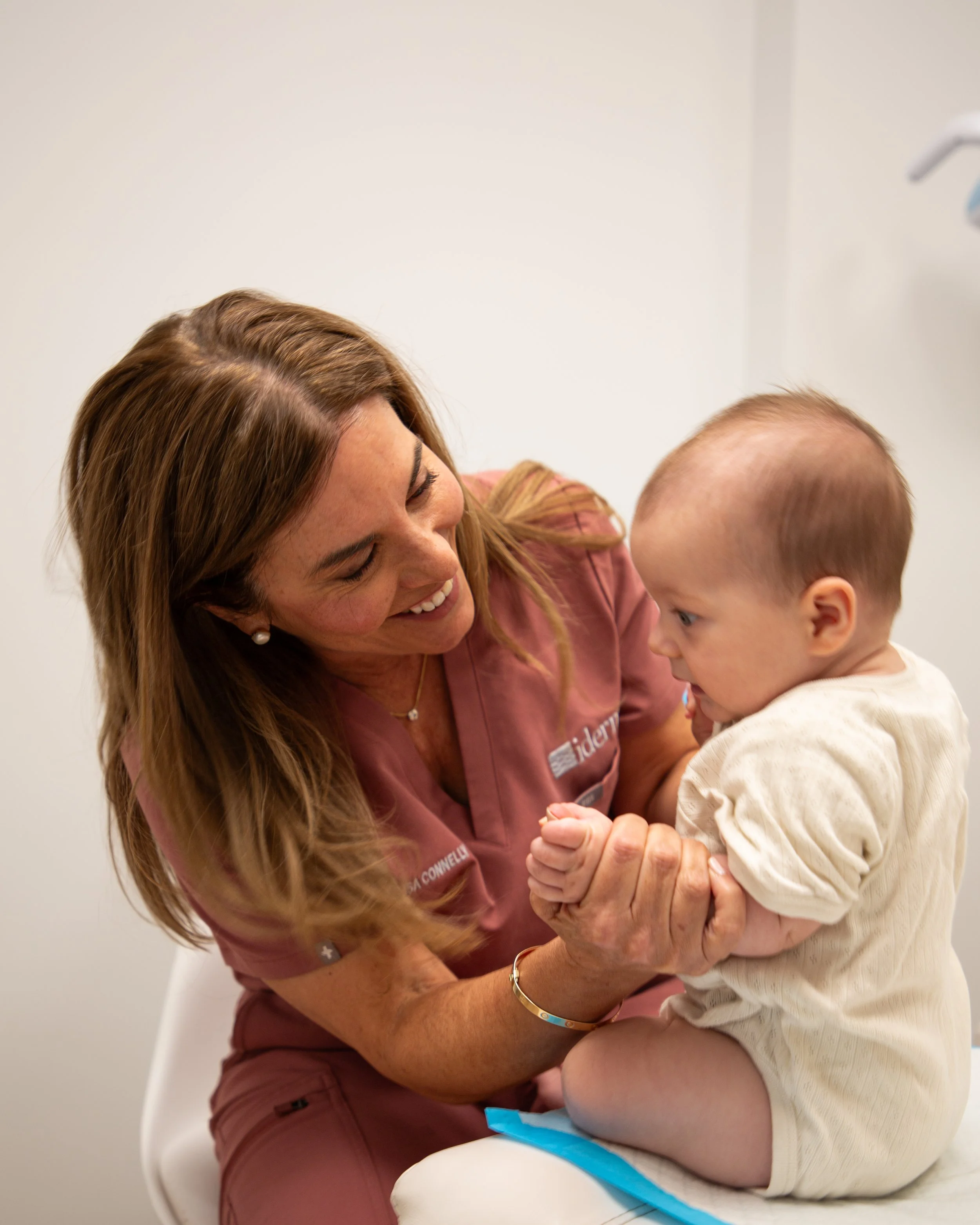A woman with long, light brown hair smiling while holding a young child with short, light hair who is looking into her eyes. They are indoors against a plain white wall.
