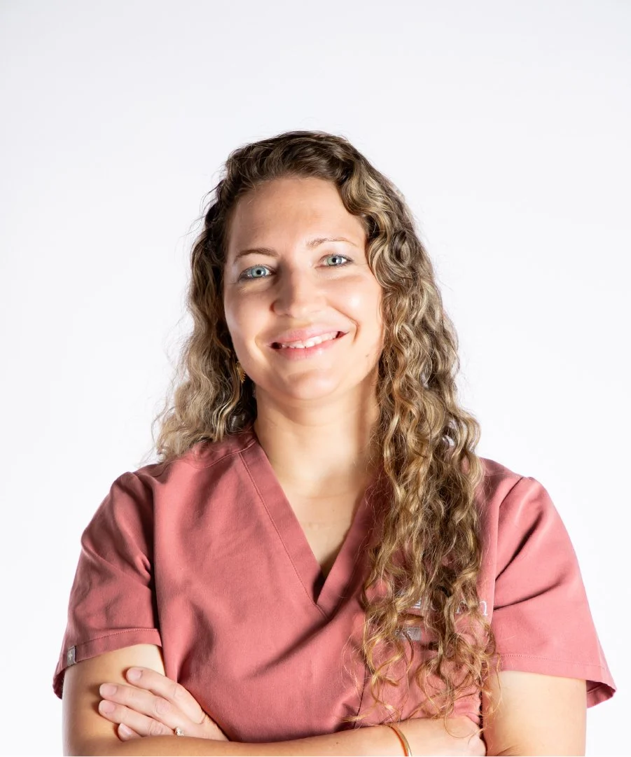 A woman with long curly hair wearing a pink scrub top, smiling with arms crossed against a plain white background.
