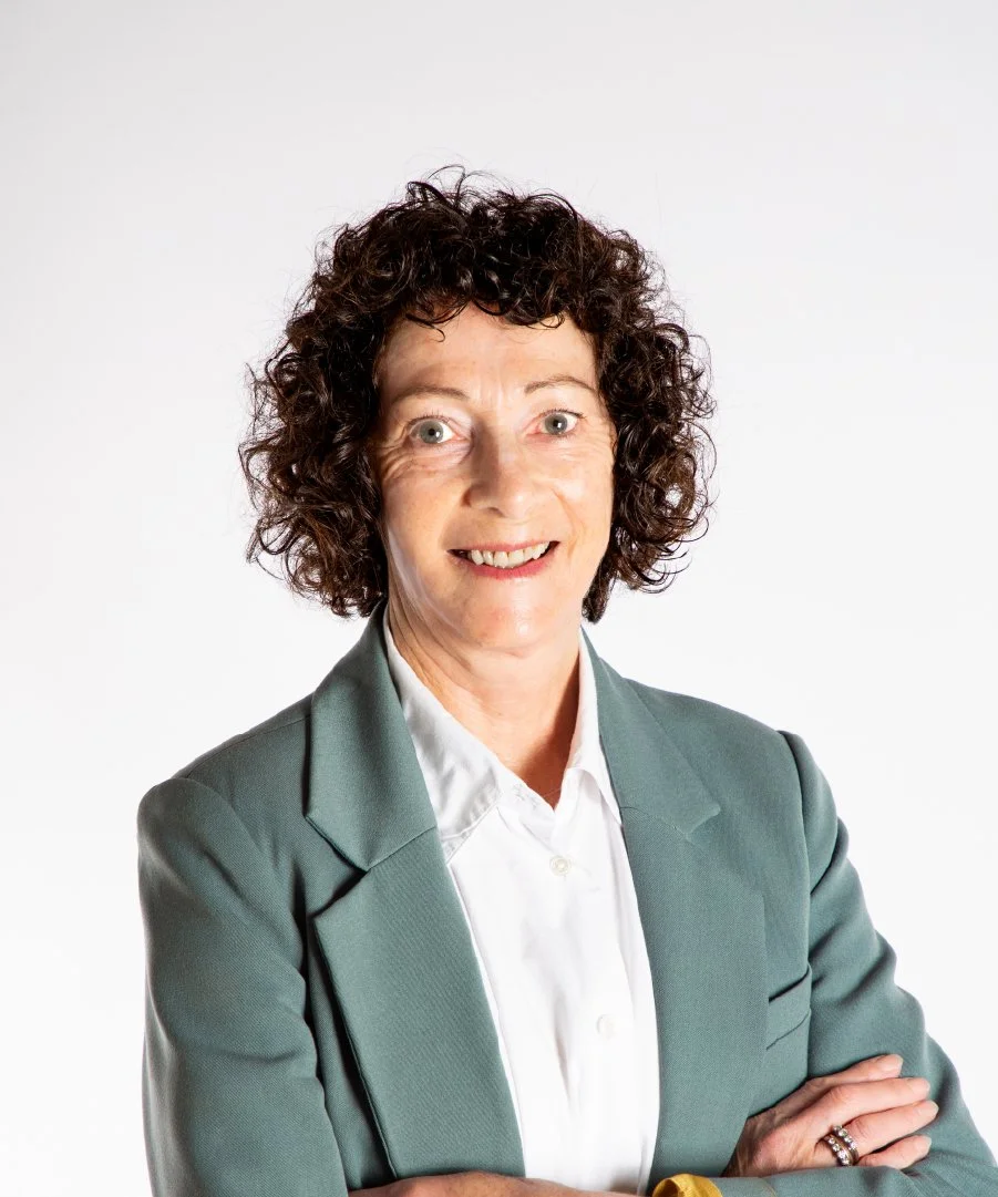 A woman with short curly brown hair wearing a gray blazer and white shirt, smiling with arms crossed, against a plain white background.