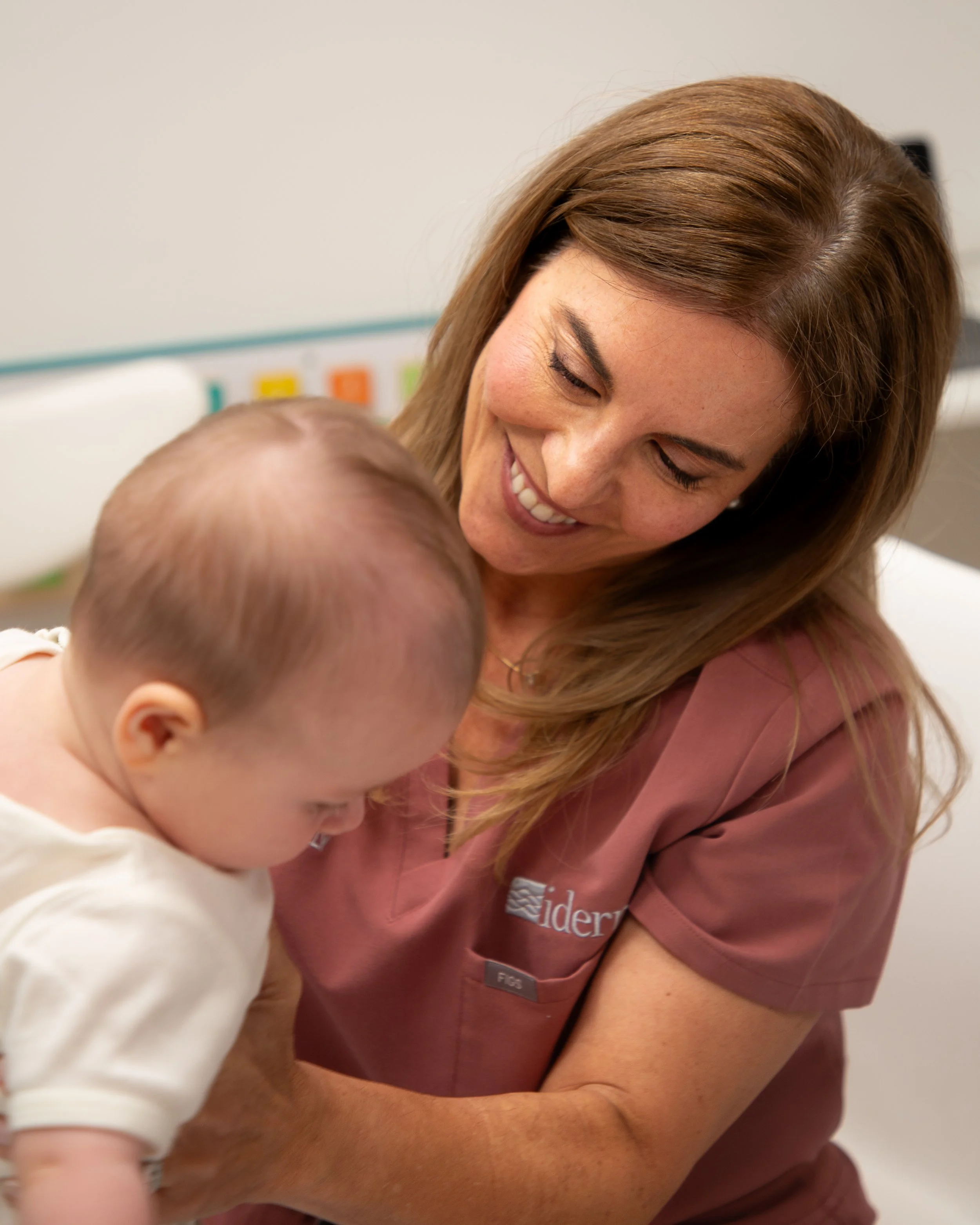 A woman with long brown hair smiling while interacting with a young child in a medical or healthcare setting.