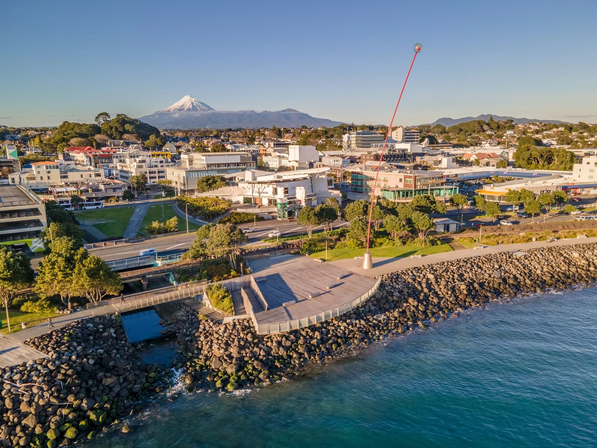 A coastal cityscape with buildings, trees, and a mountain with a snow-capped peak in the background, along with a large weather monitoring antenna near the shoreline.