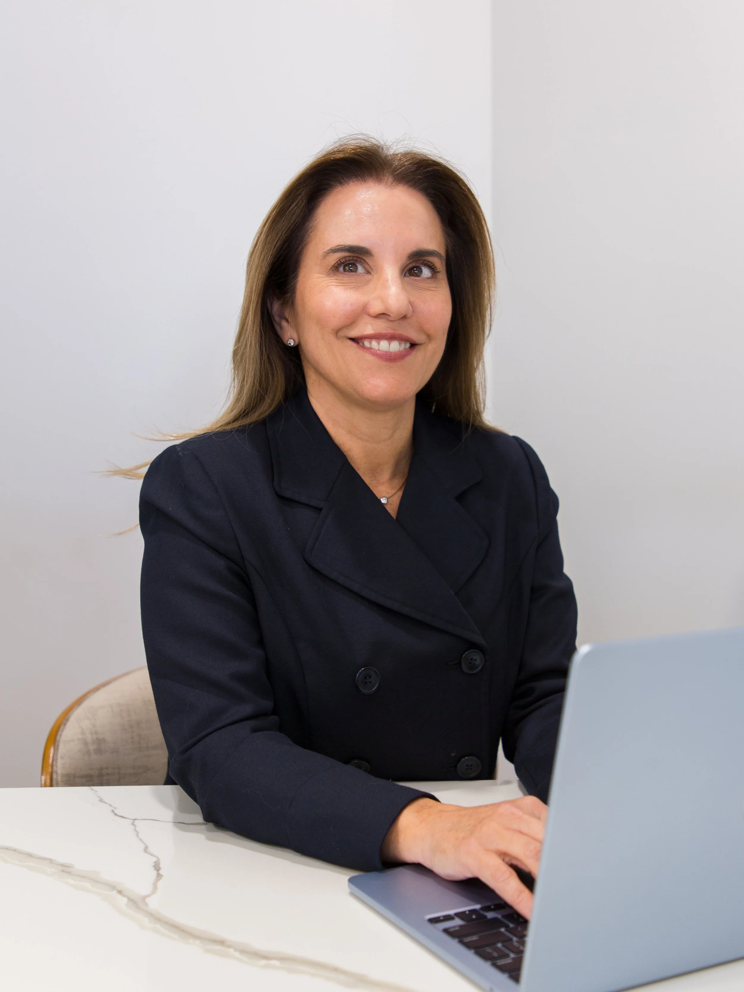 A woman with brown hair and a dark blazer working on a laptop at a white marble table.