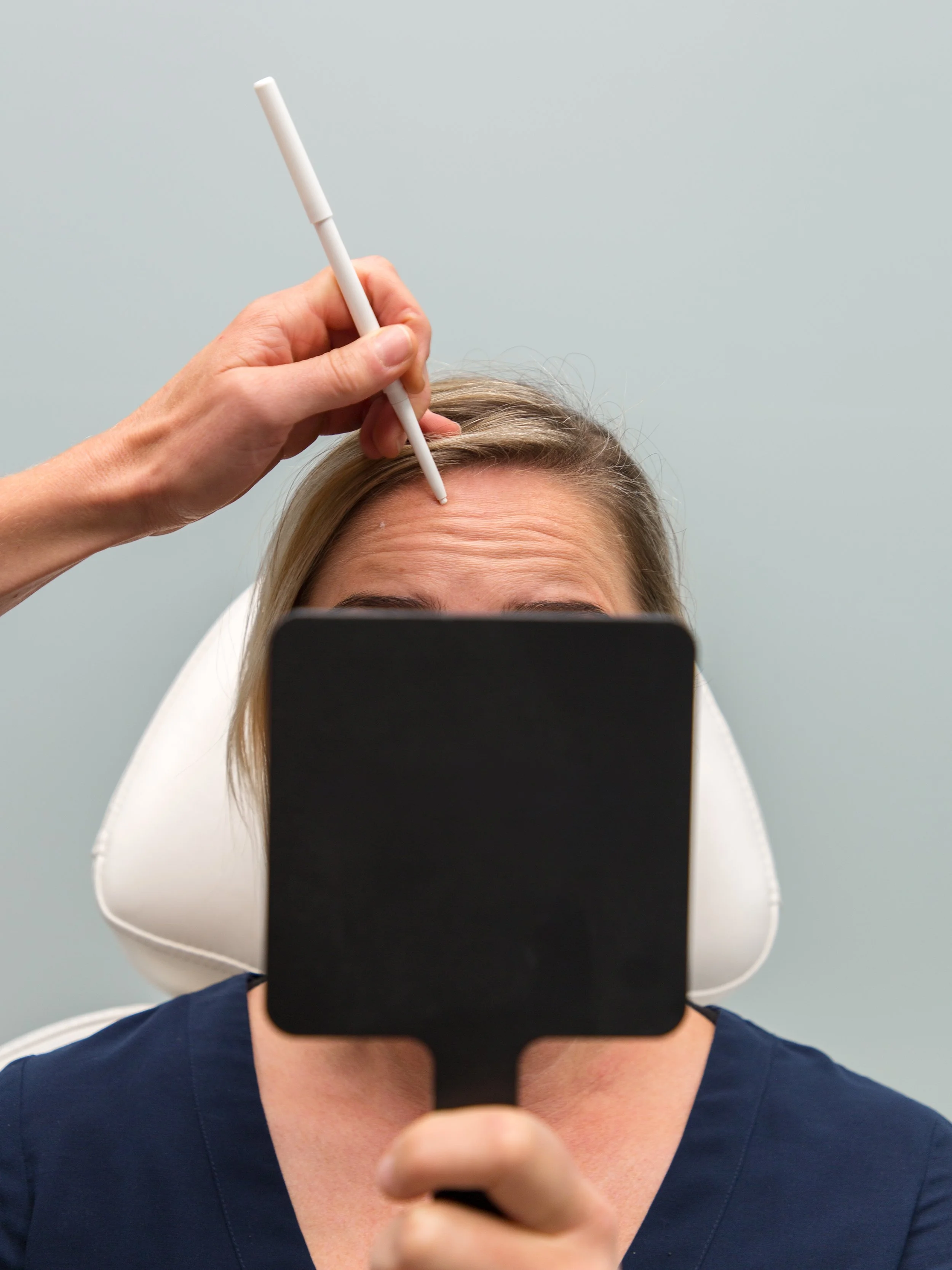 A person taking a selfie in a mirror with a smartphone, showing their forehead and part of their nose, with a hand holding a white stylus near their forehead, against a plain light blue wall background.