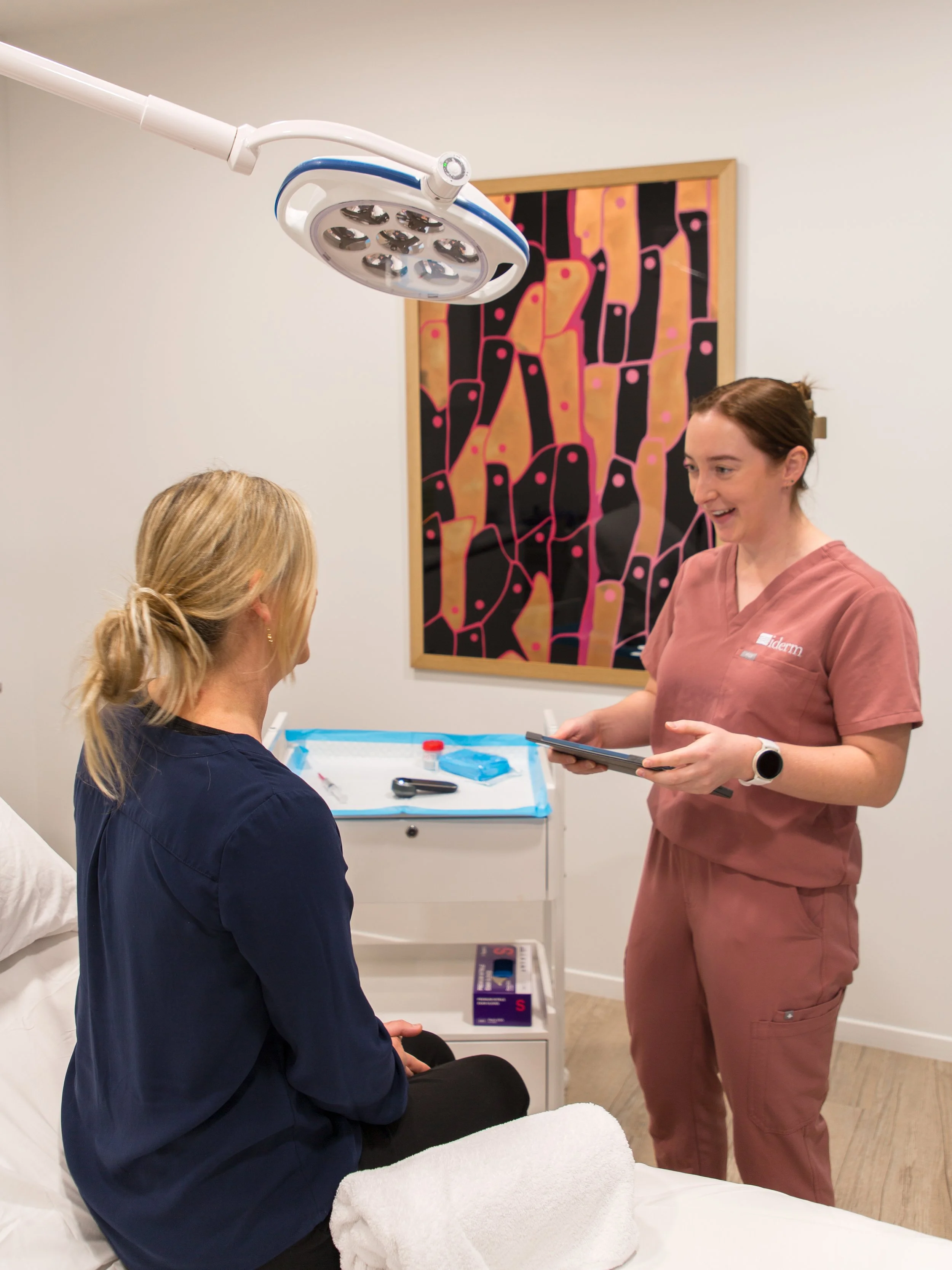 A woman in medical scrubs talking to a patient sitting on a bed, with a white medical monitor and a colorful abstract painting on the wall behind them.