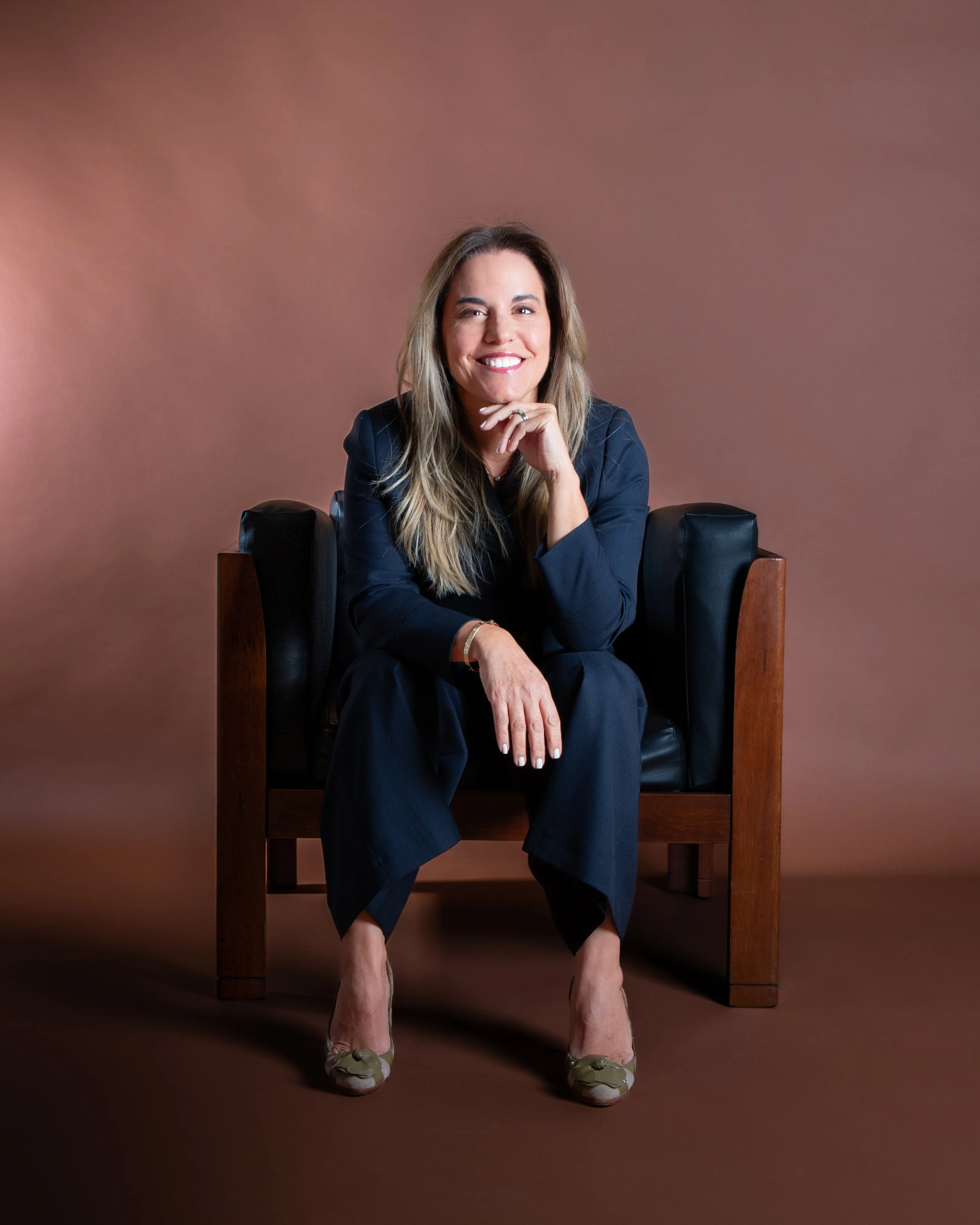 Smiling woman with long blond hair sitting on a wooden and leather chair, wearing a blue blazer and matching pants, beige heels, and a ring, against a plain brown background.