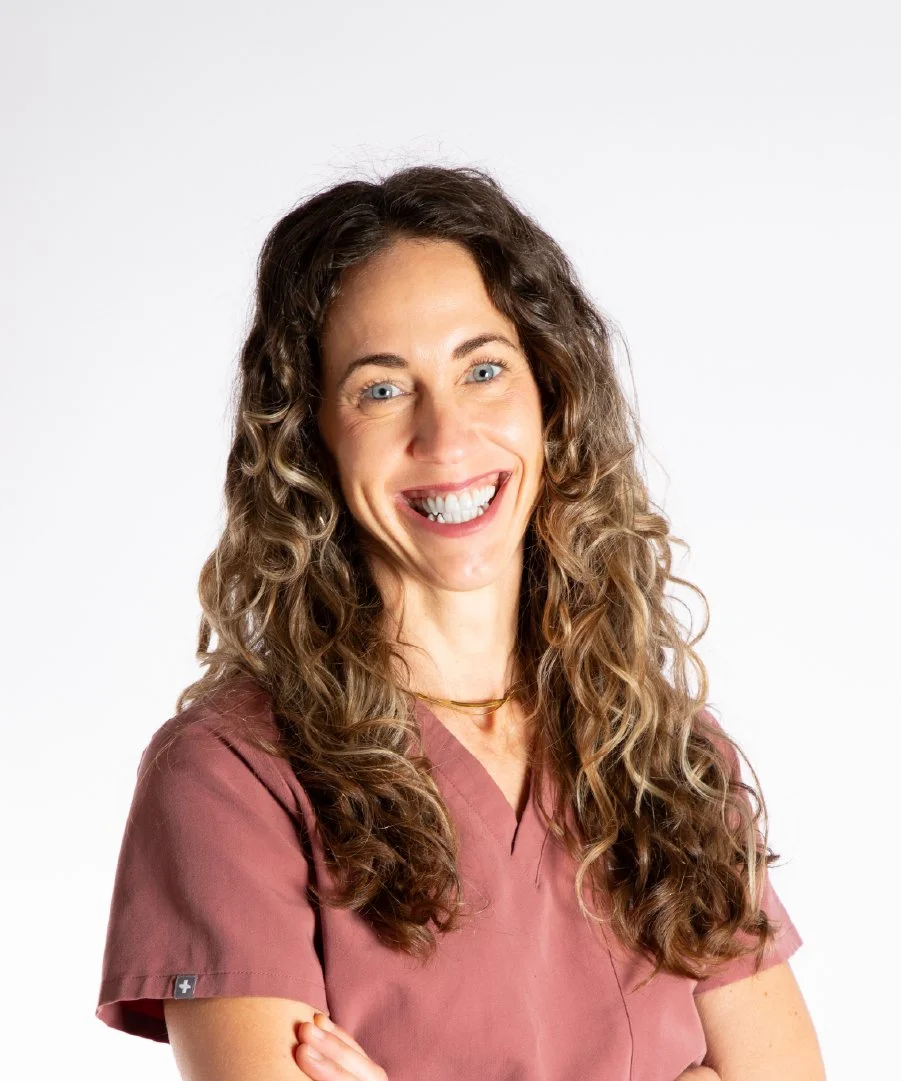 A woman with curly brown hair smiling and crossing her arms, wearing a mauve scrub top with a small cross tag on the sleeve, against a plain white background.