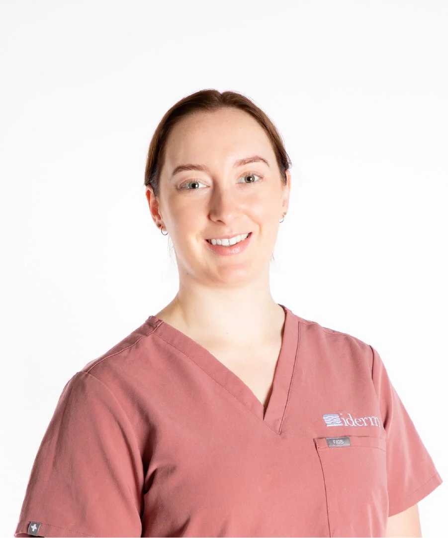 A woman with light skin and reddish-brown hair smiling, wearing a pink medical scrub top with a logo and name tag, standing against a white background.